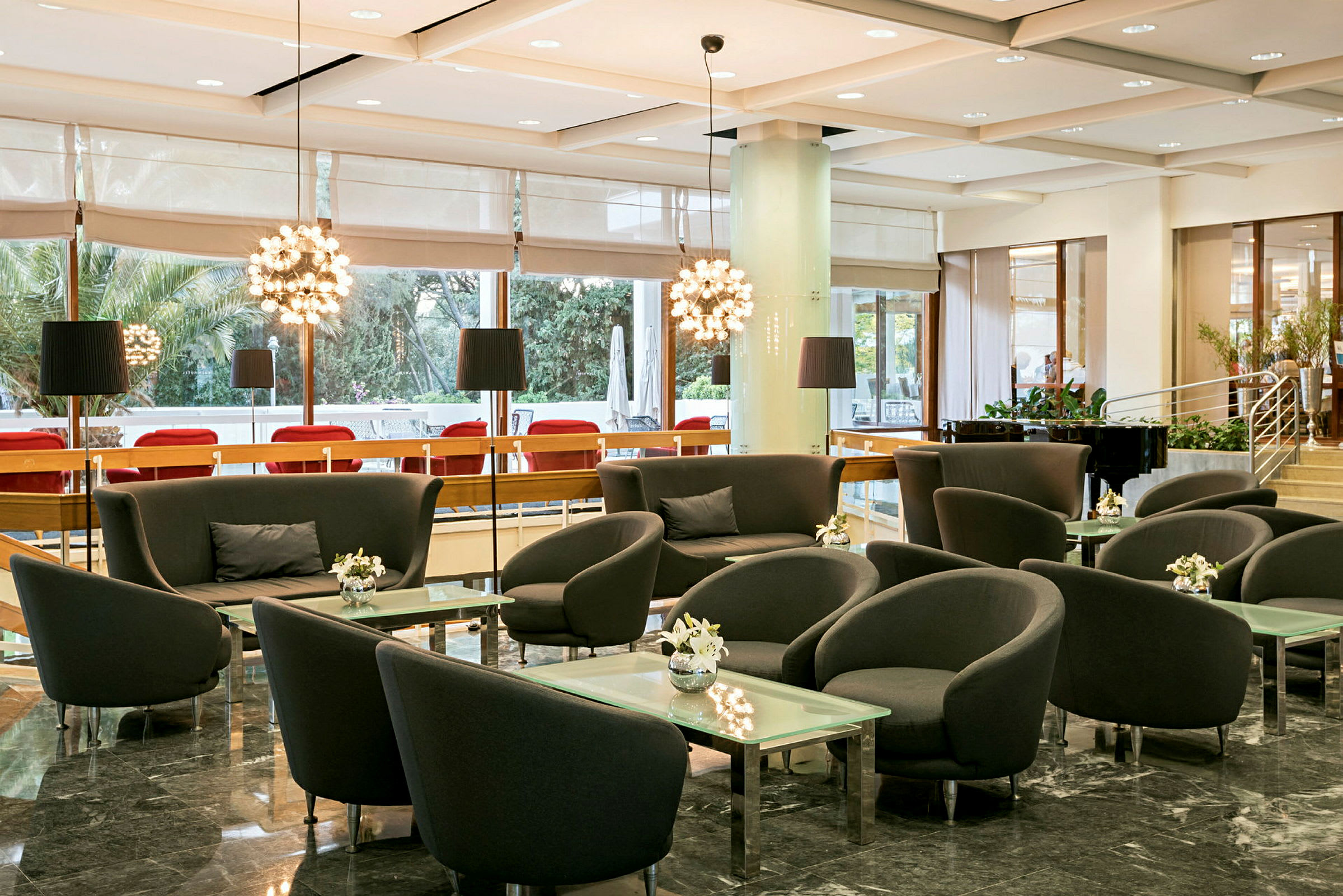 En velbelyst lounge med siddegrupper bestående af lænestole og sofaer omkring glasborde, omgivet af store vinduer og moderne indretning. (AI-genereret tekst)