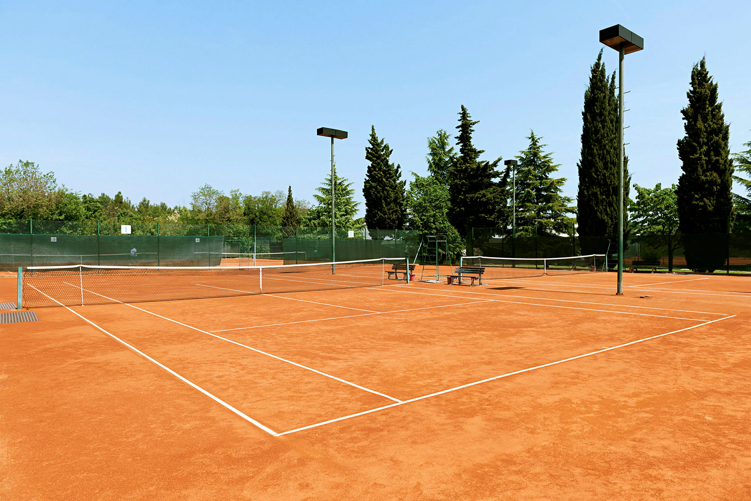 En tennisbane med grusunderlag omgivet af træer og hegn. (AI-genereret tekst)