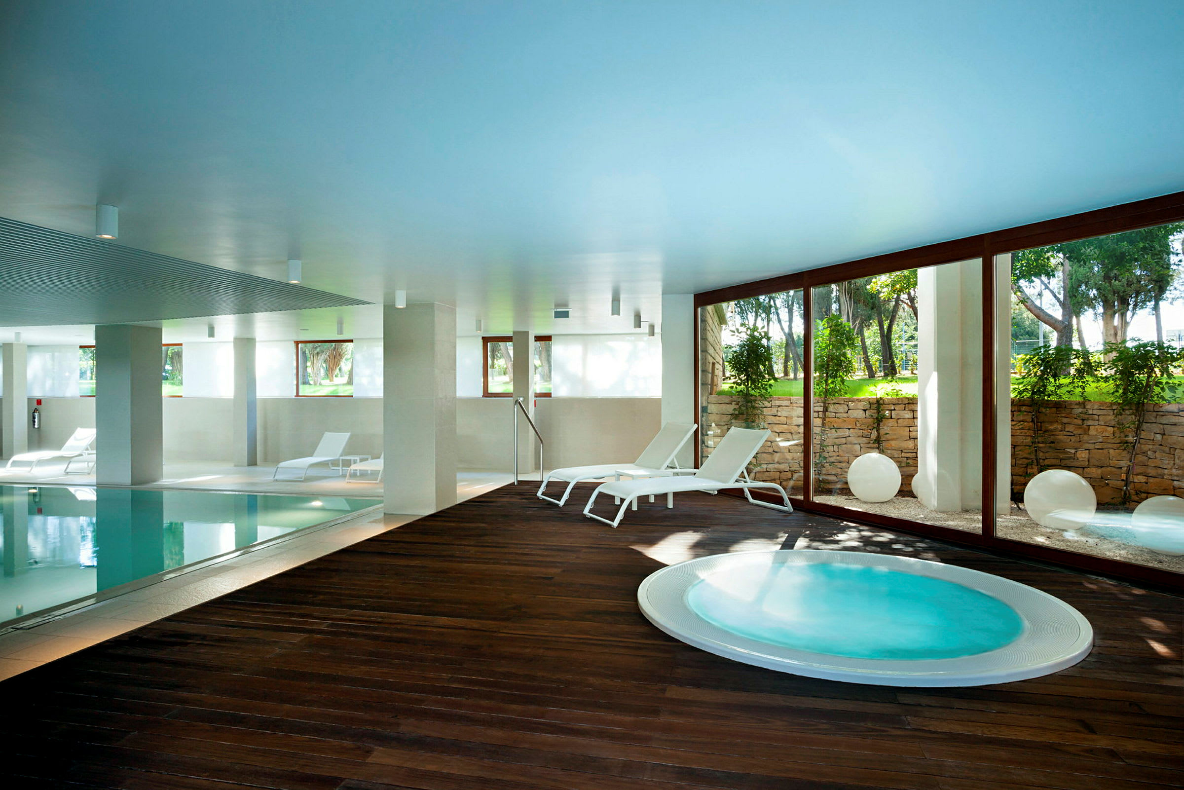 Indendørs pool med liggestole og jacuzzi ved siden af, samt glasvæg med udsigt til en frodig have. (AI-genereret tekst)
