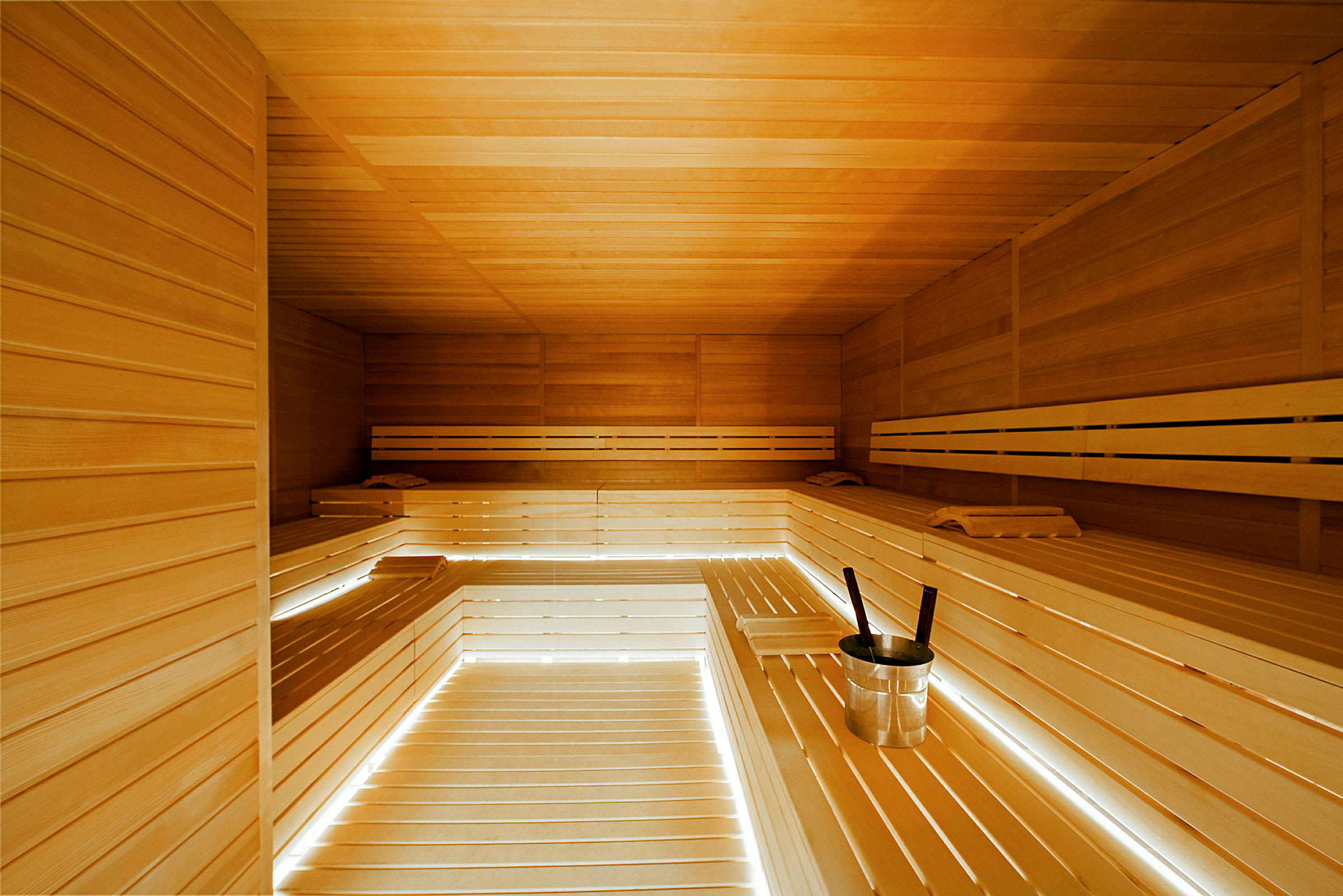 Billedet viser interiøret i en sauna med lyse træpaneler og bænke. En spand med øse står på en af bænkene. (AI-genereret tekst)