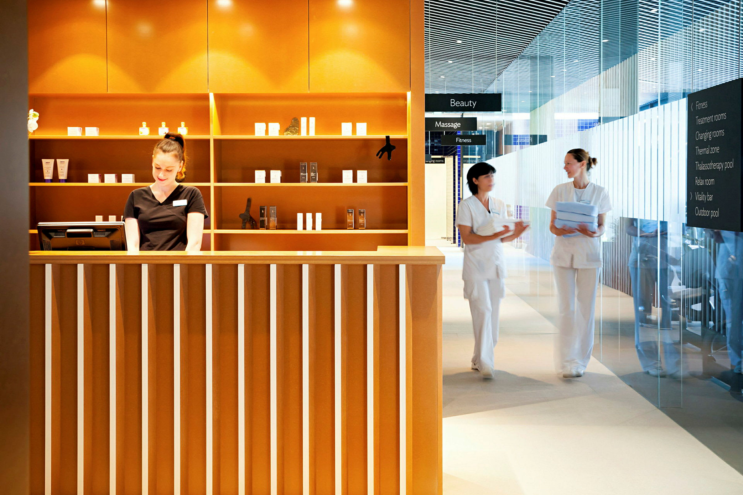 Receptionen for en spa- eller skønhedsfacilitet med hylder fyldt med produkter og to personer i arbejdstøj samtaler ved siden af. (AI-genereret tekst)