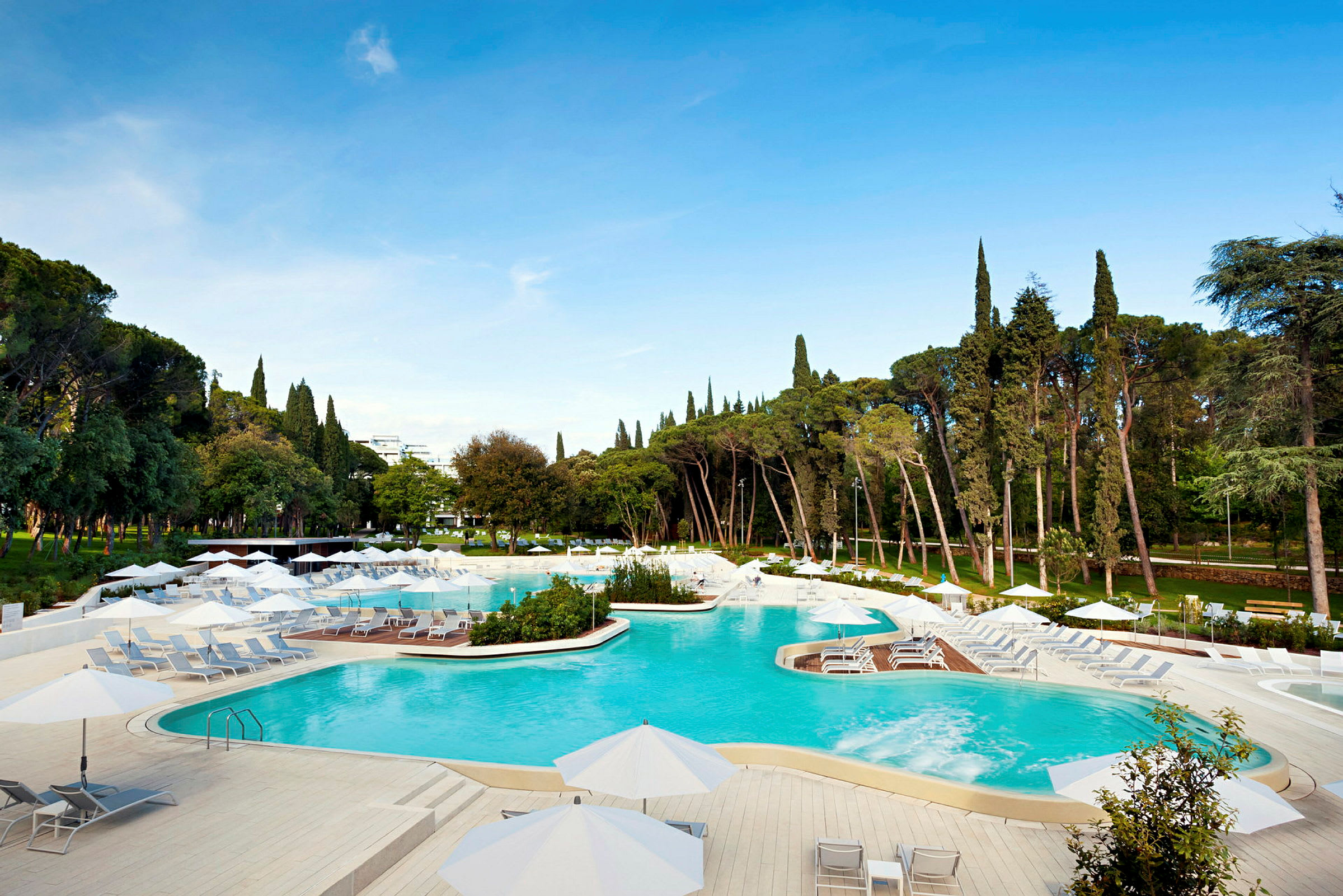 En lys og moderne pool med parasoller og liggestole, omgivet af grønne træer og en klar blå himmel. (AI-genereret tekst)