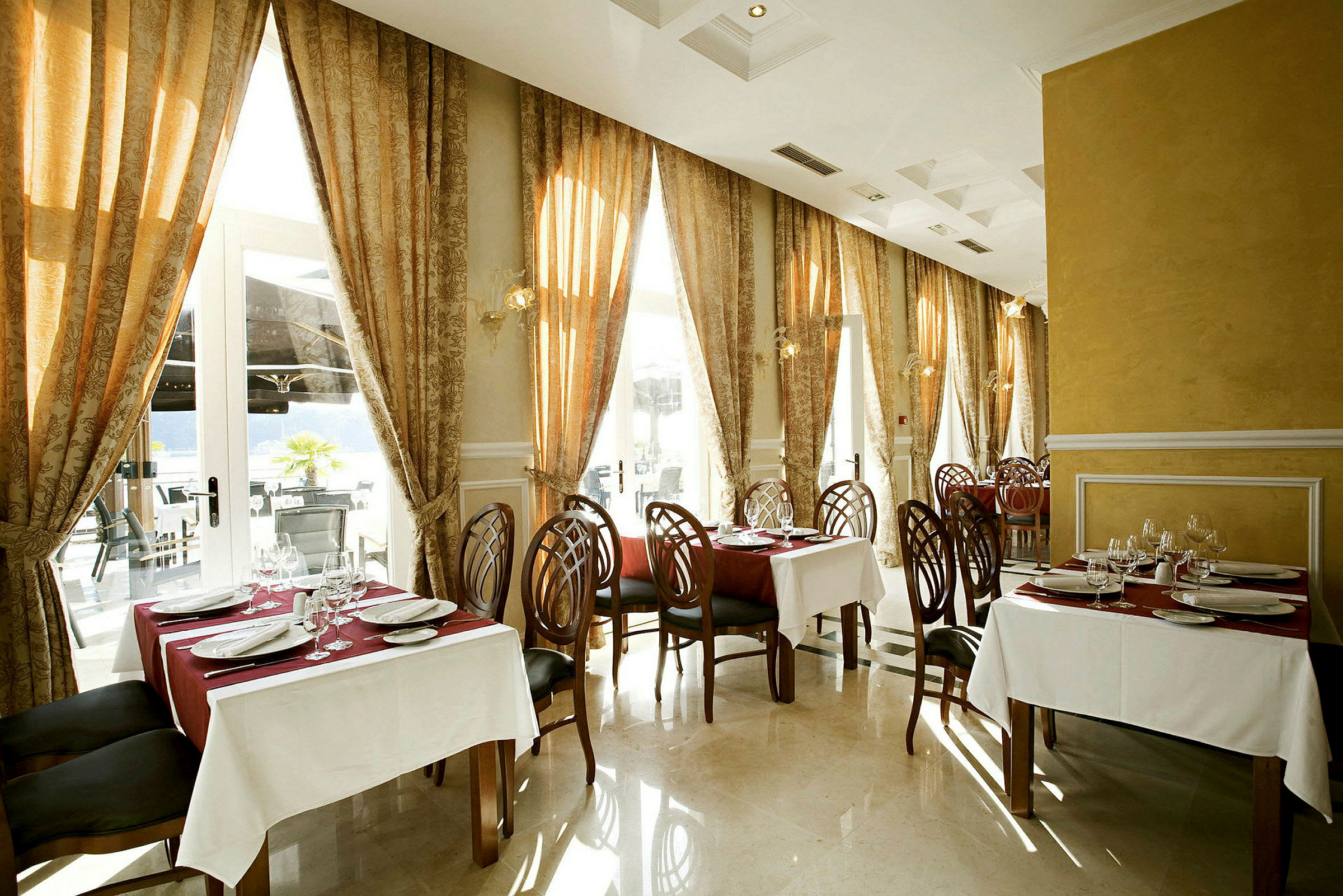Et elegant restaurantområde med borde dækket med hvide duge og røde servietter, omgivet af dekorerede stole og store vinduer med tunge gardiner. (AI-genereret tekst)
