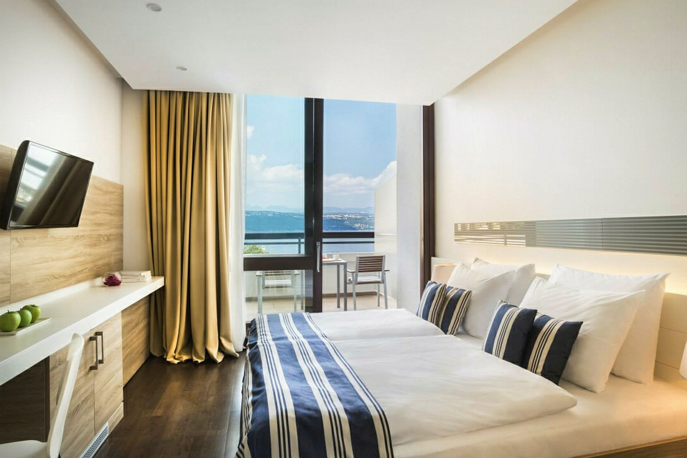 Et hotelværelse med en dobbeltseng, sengetøj i blå- og hvidstribet mønster og en stor glasvæg, der viser en balkon med udsigt over havet. Værelset har også et TV og skrivebord. (AI-genereret tekst)