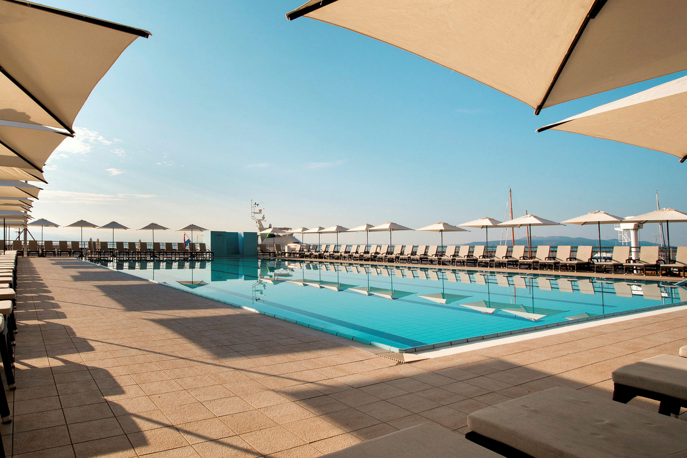 En swimmingpool med liggestole og parasoller omkring, placeret udendørs under en blå himmel. (AI-genereret tekst)