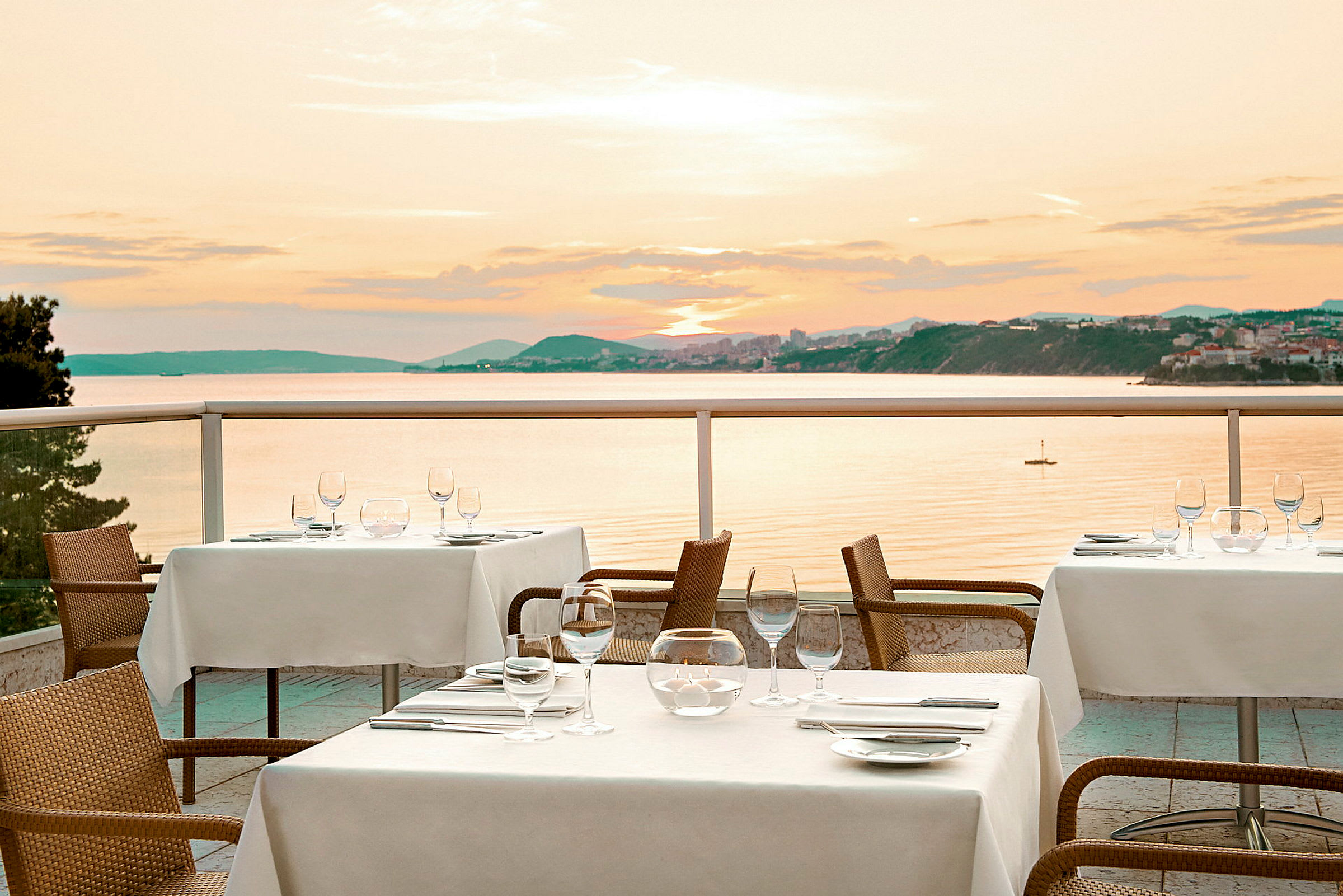Borde med hvide duge på en terrasse med udsigt over havet og solnedgangen. (AI-genereret tekst)