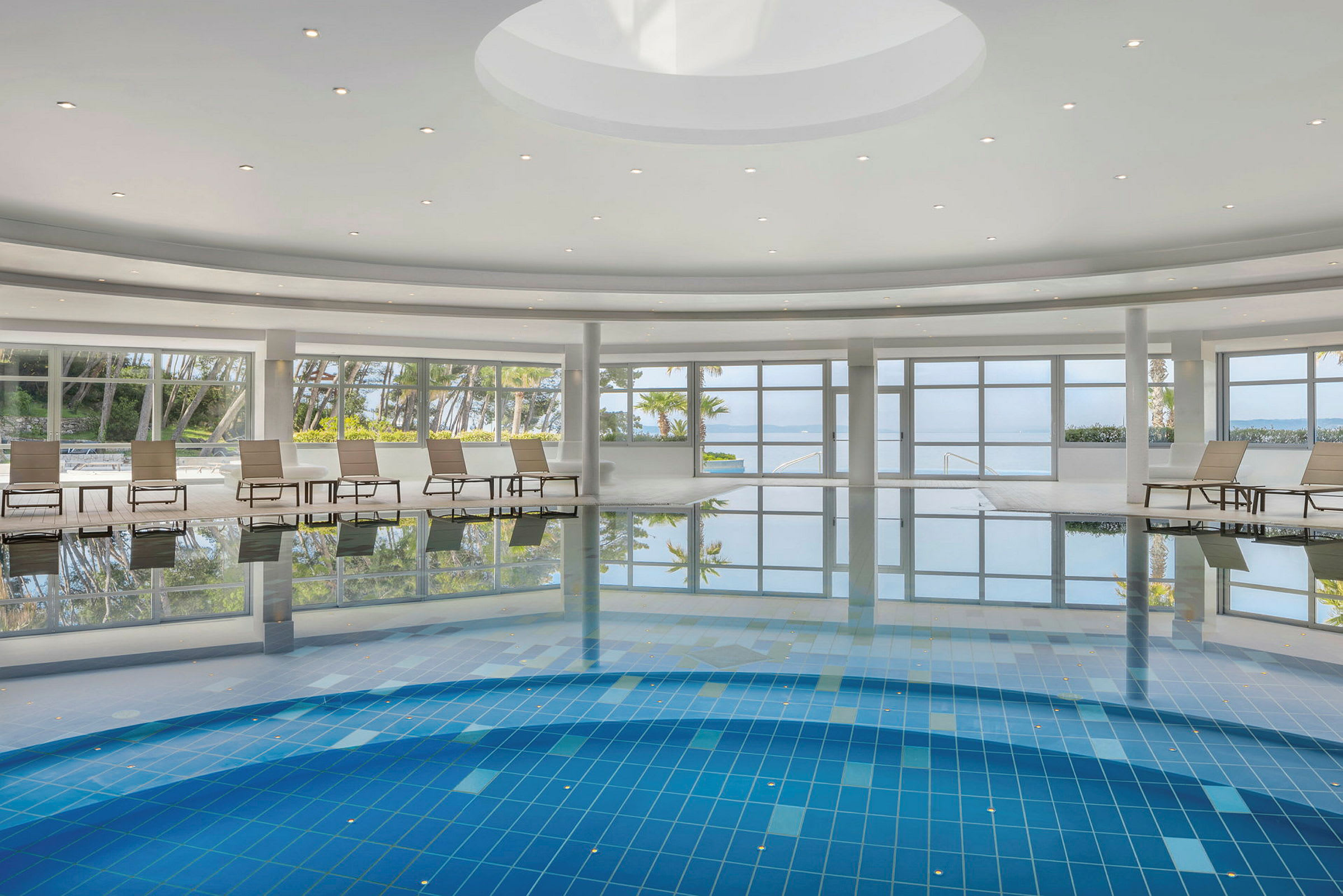 Indendørs swimmingpool med liggestole og store vinduer med udsigt over omgivelserne. (AI-genereret tekst)