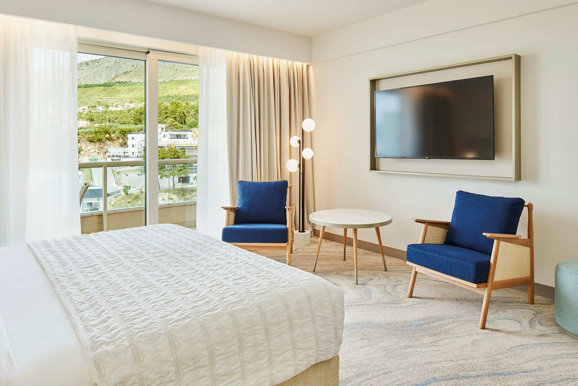 Hotelværelse med seng, to blå stole, et rundt bord og et vægmonteret TV. Altan og grøn udsigt ses gennem glasdøren. (AI-genereret tekst)