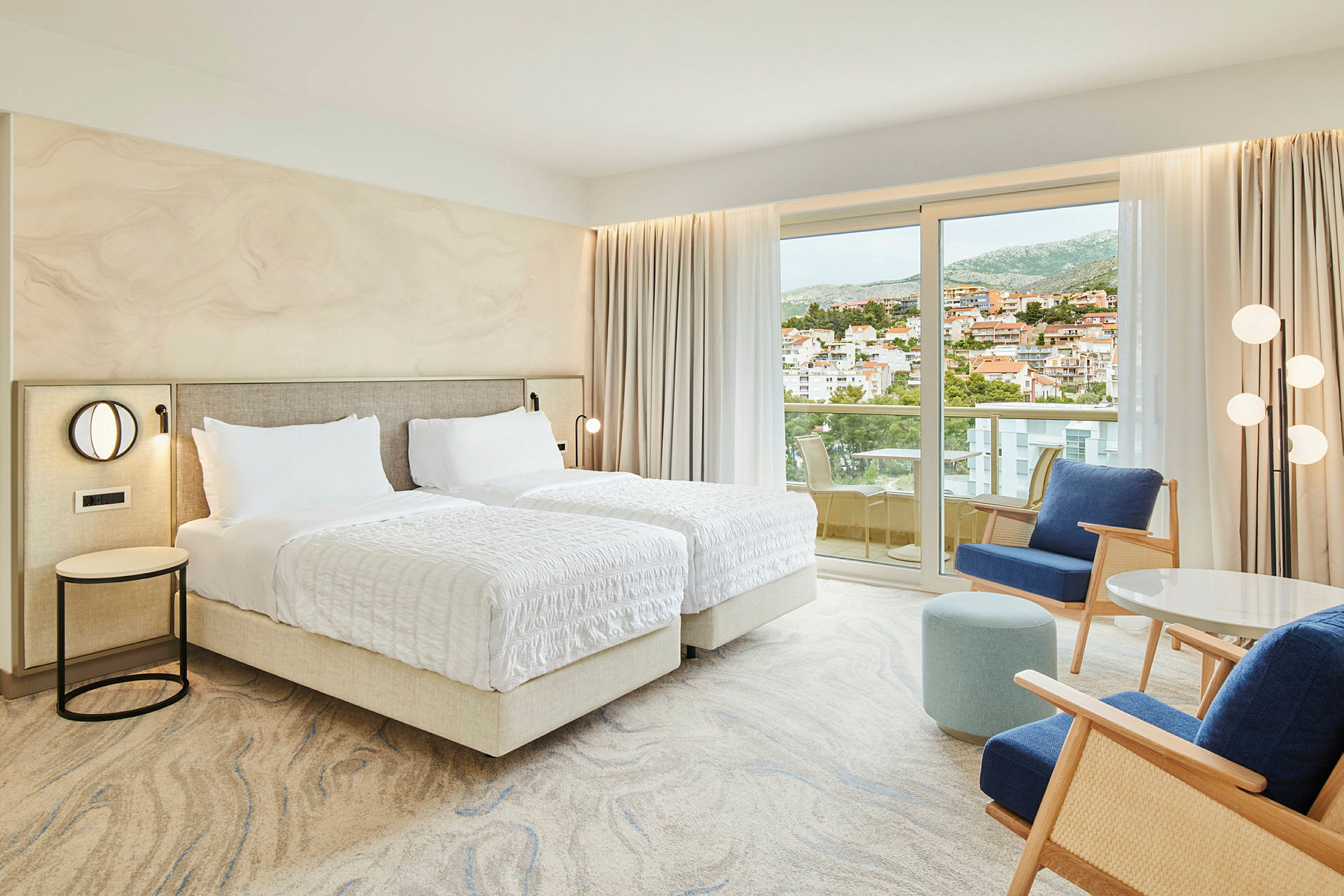 Hotelværelse med to enkeltsenge, siddeområde og balkon med udsigt over en by. (AI-genereret tekst)