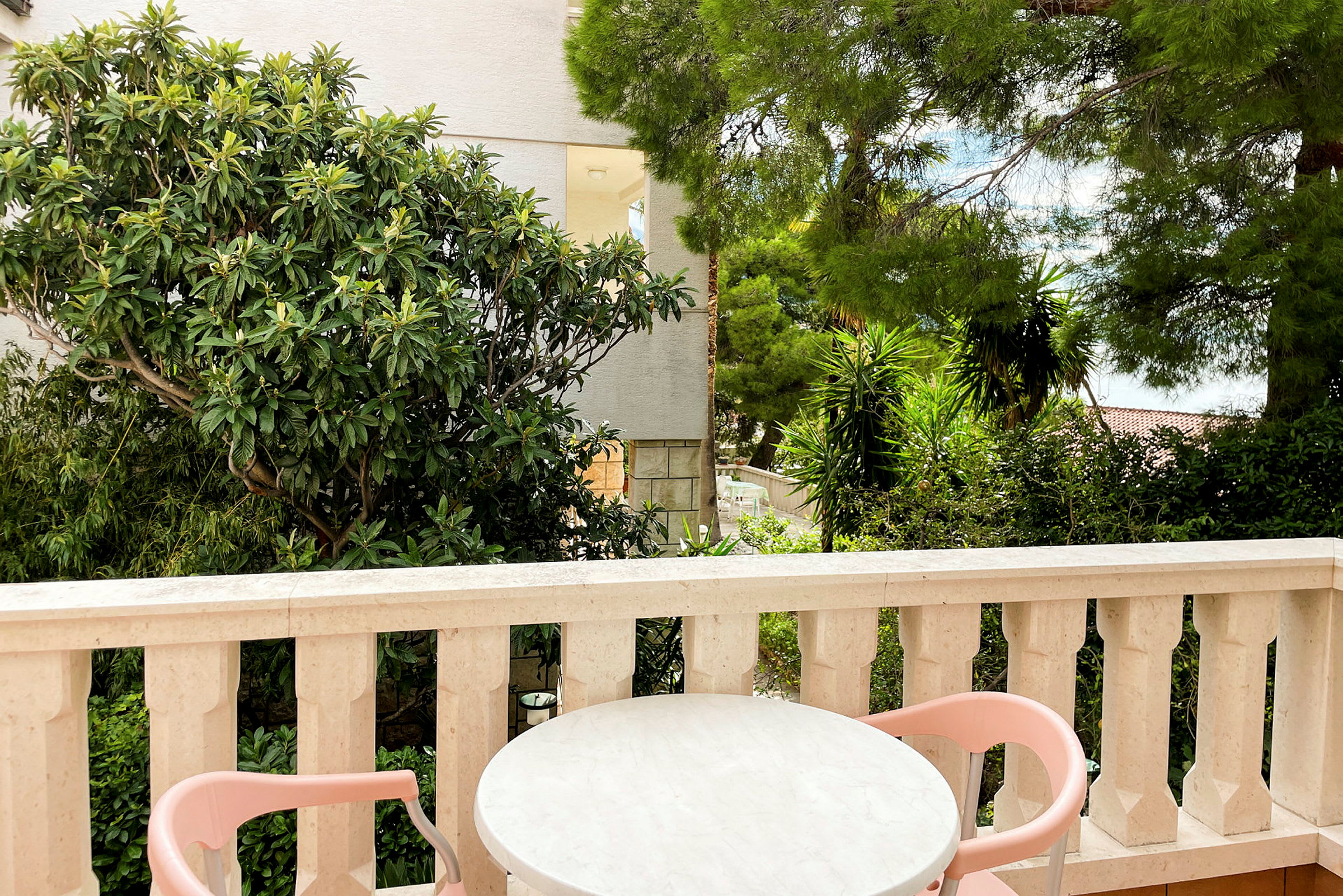 En terrasse med et bord og to lyserøde stole, omgivet af grønne planter og træer. (AI-genereret tekst)