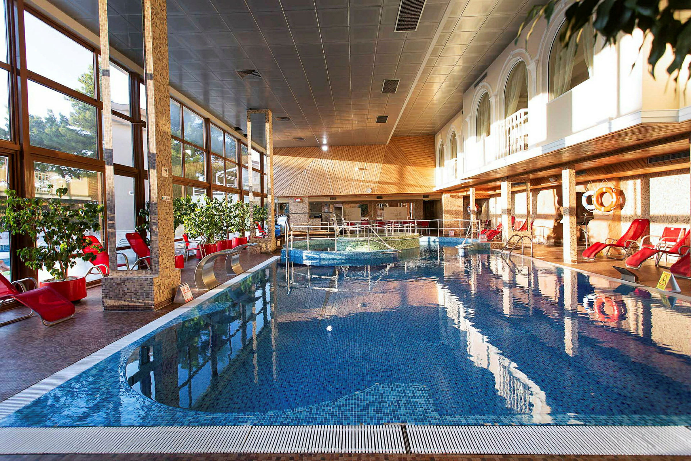 En indendørs pool med røde liggestole omkring, planter og store vinduer. (AI-genereret tekst)