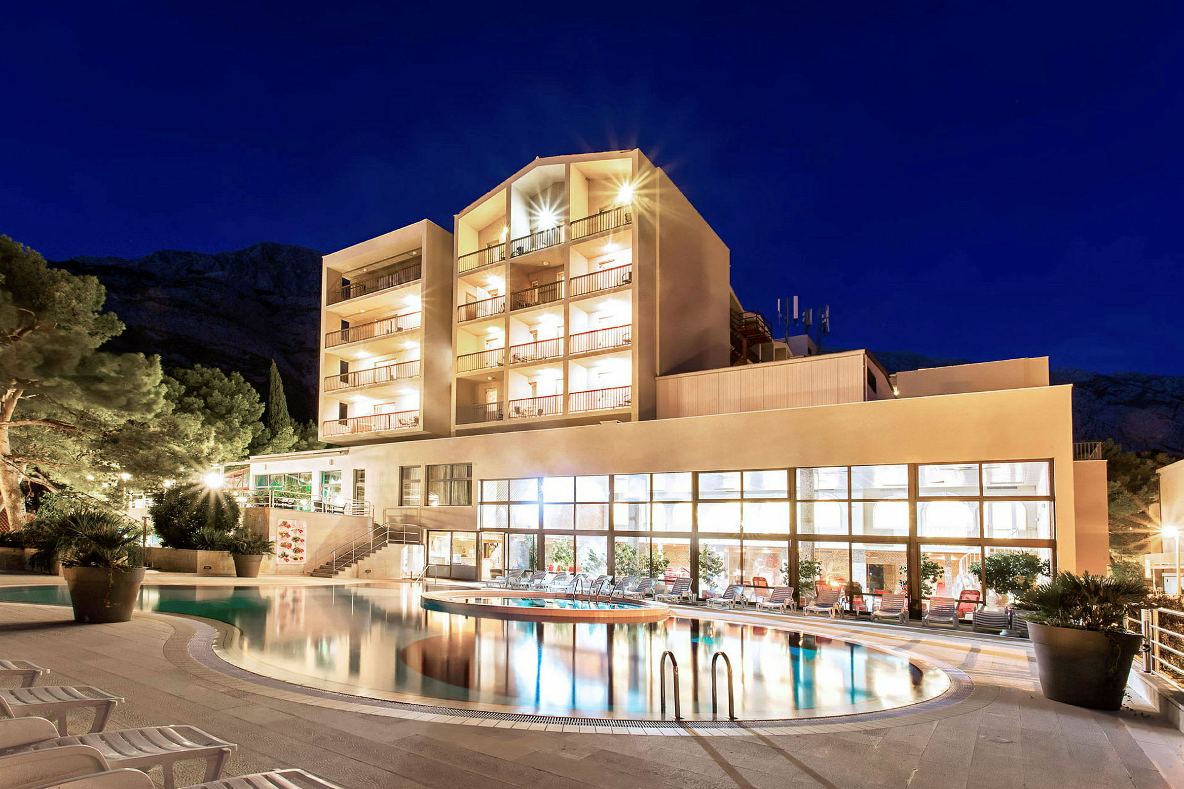 Et hotel med en oplyst facade ved en swimmingpool om natten. (AI-genereret tekst)