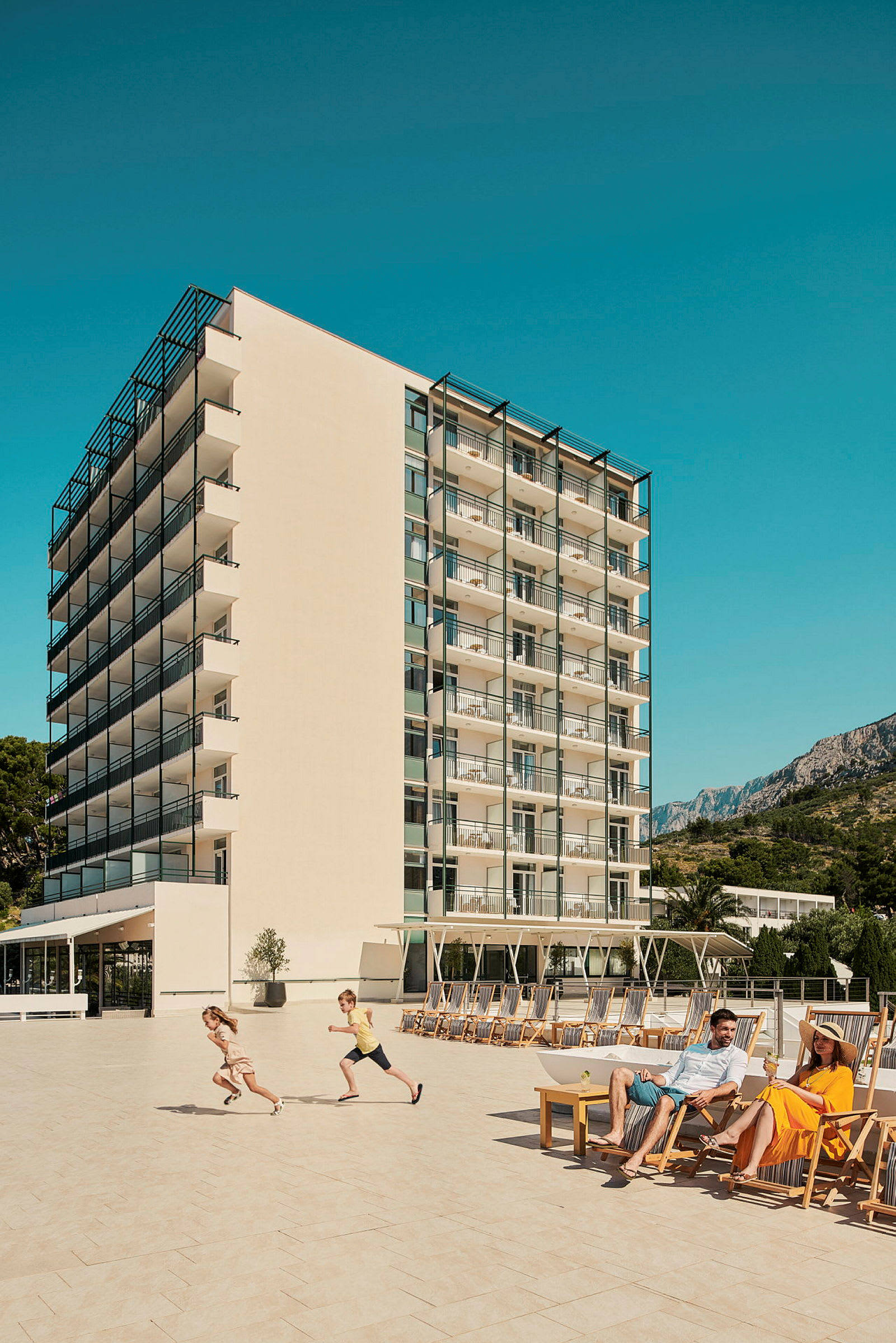 Billede af et hotel med altaner i flere etager, en stor terrasse med solsenge, og personer der nyder solen. (AI-genereret tekst)