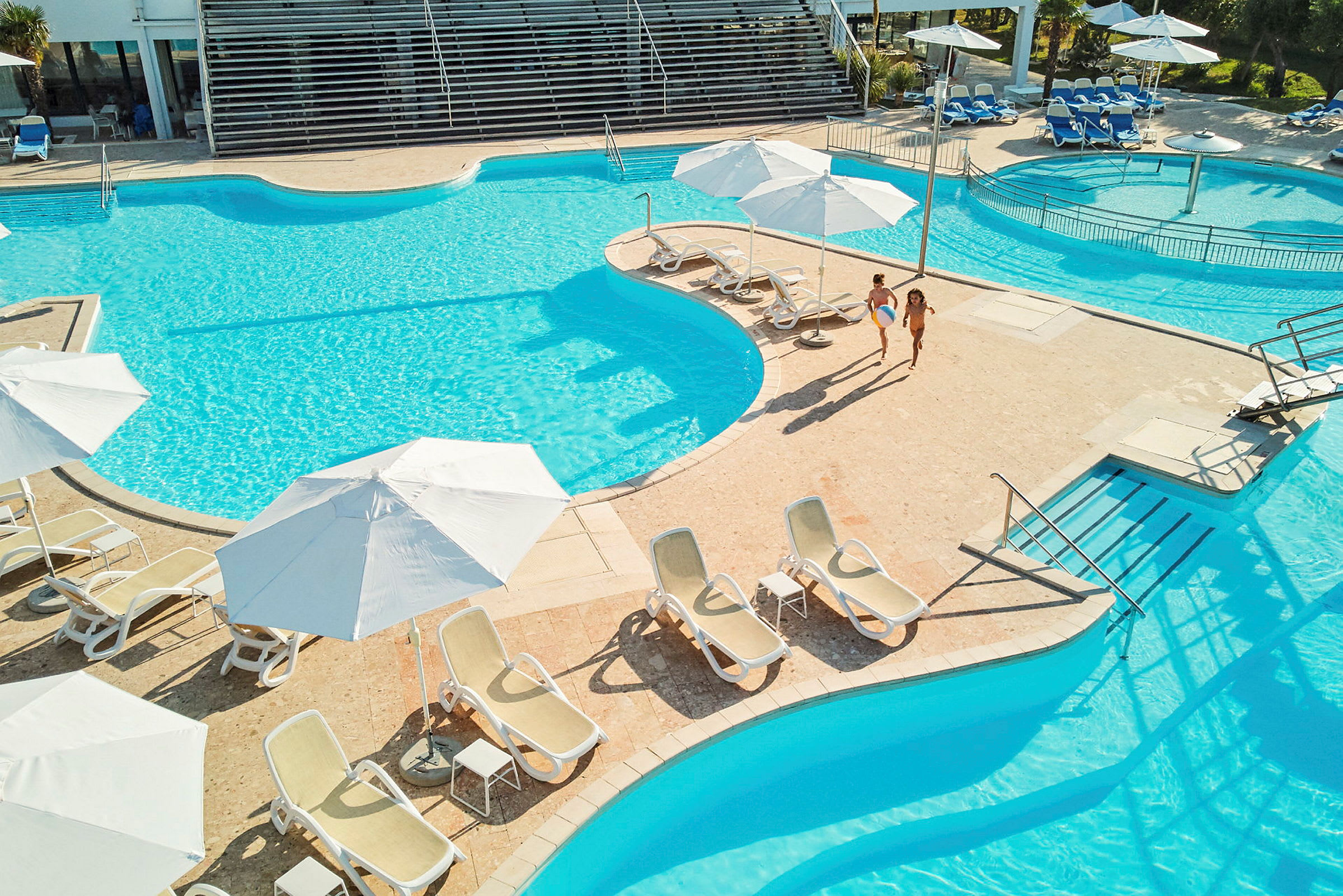 To personer går ved en pool omgivet af liggestole og parasoller. (AI-genereret tekst)