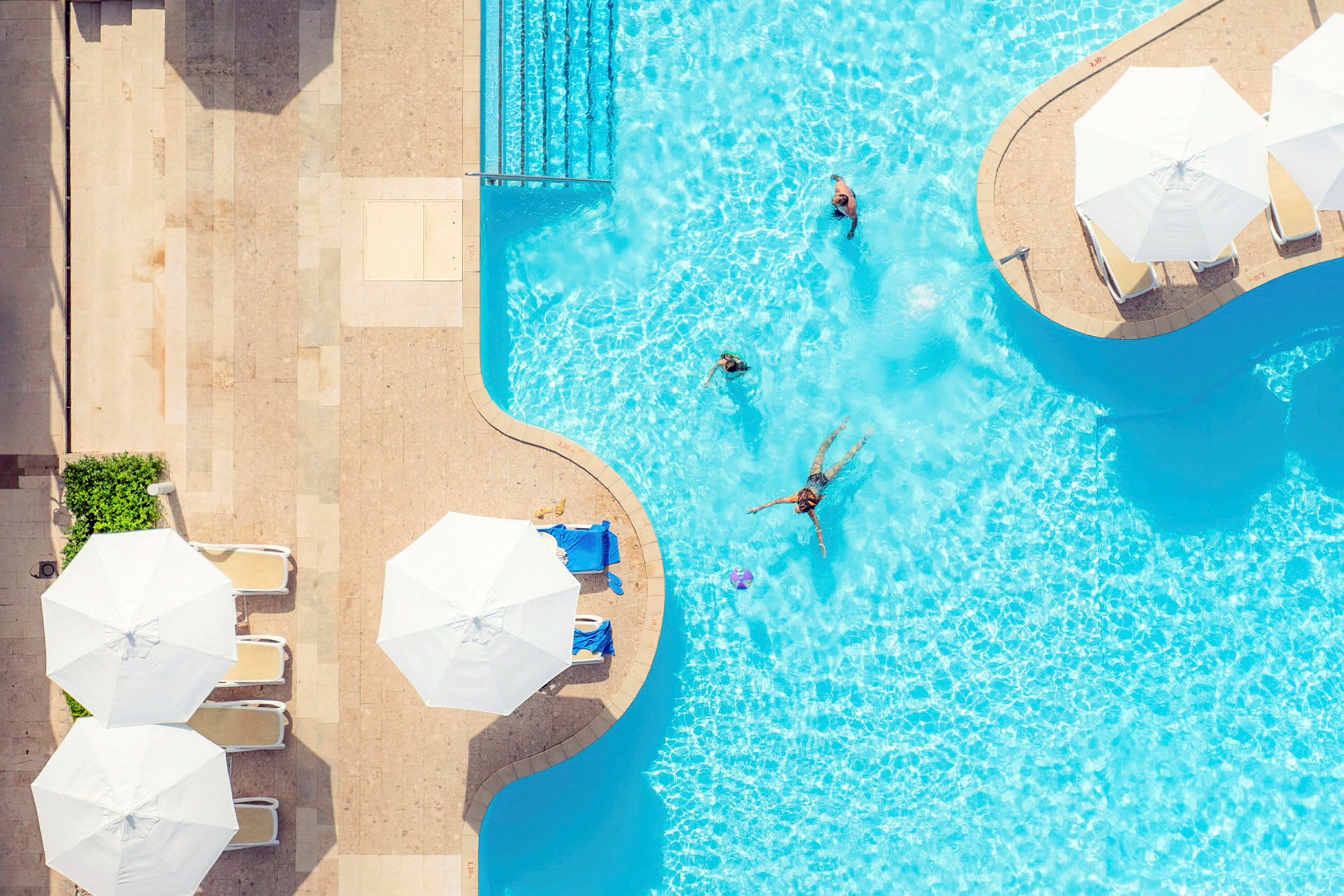 Udsigt oppefra af en pool med nogle mennesker, der svømmer, og liggestole under parasoller ved poolkanten. (AI-genereret tekst)