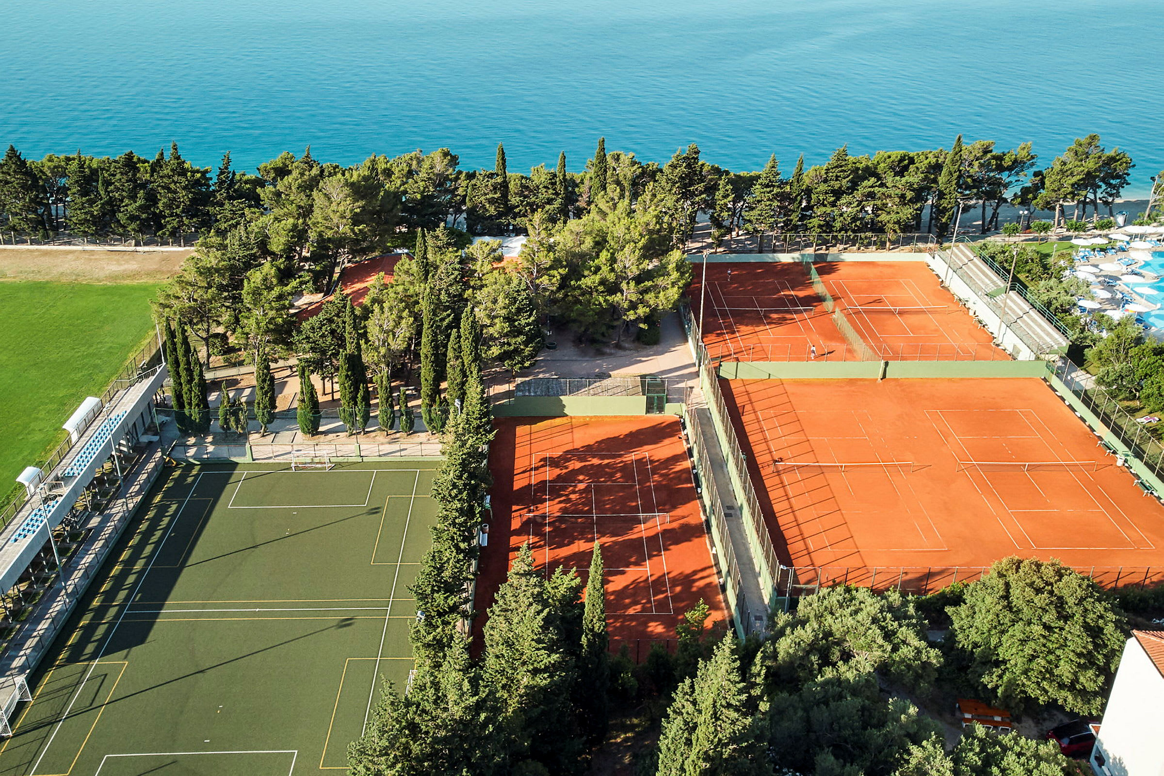 Luftfoto af sportsfaciliteter med tennisbaner og fodboldbane, omgivet af træer og hav. (AI-genereret tekst)
