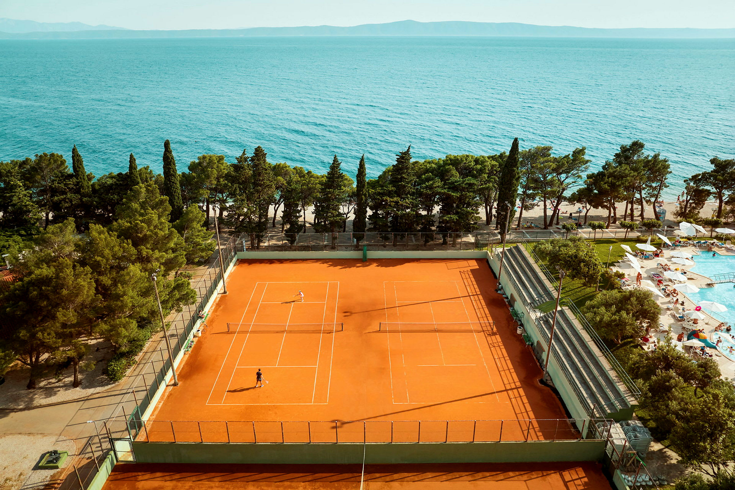 Tennisbane med spillere tæt på havet og et poolområde. (AI-genereret tekst)