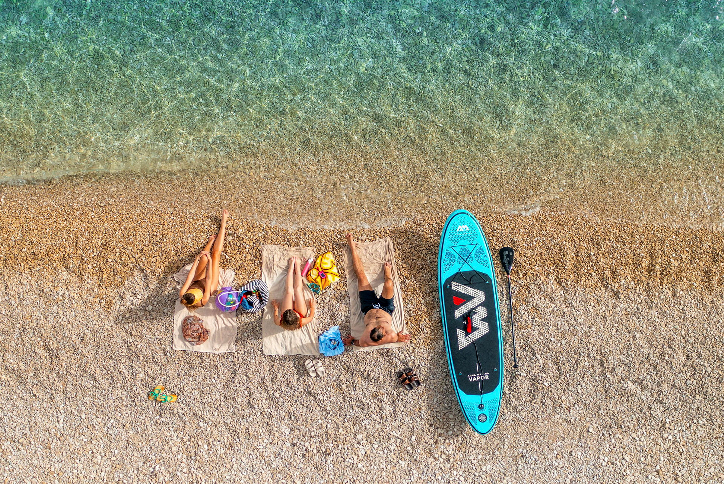 Tre personer ligger på håndklæder på stranden ved siden af et paddleboard. (AI-genereret tekst)