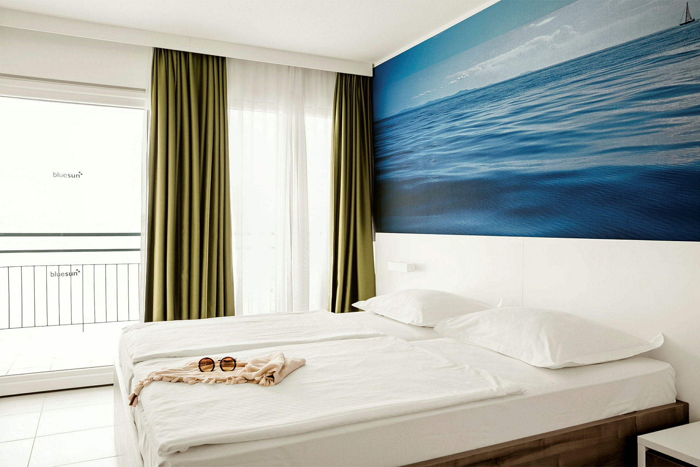 Et hotelværelse med en dobbeltseng, grønne gardiner, en hvid væg med et blåt billede af havet og et par solbriller på sengen. (AI-genereret tekst)