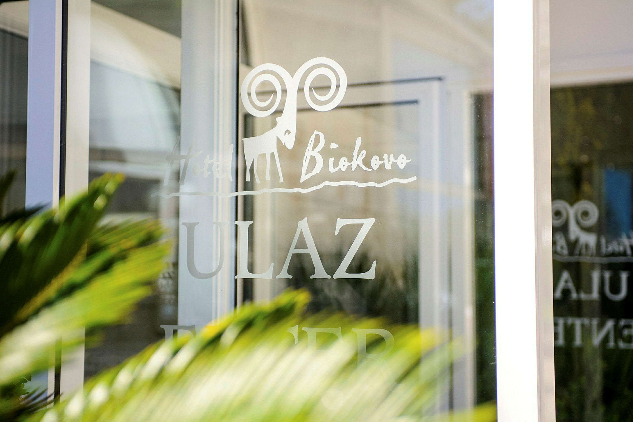 Tekst på en glasdør med ordene "Hotel Biokovo" og "ULAZ," med et tegnet symbol af en stenbuk. (AI-genereret tekst)