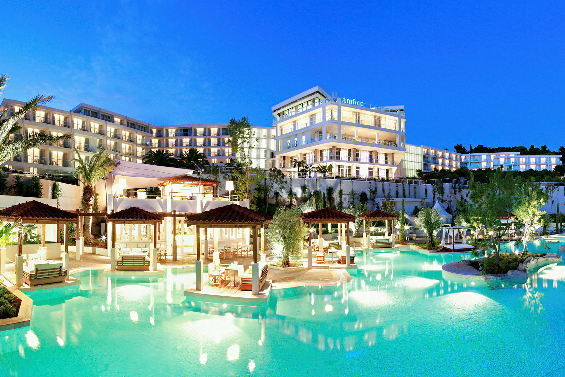 Et oplyst hotelkompleks med flere etager ved en pool, omgivet af haver og palmer, i skumringen. (AI-genereret tekst)