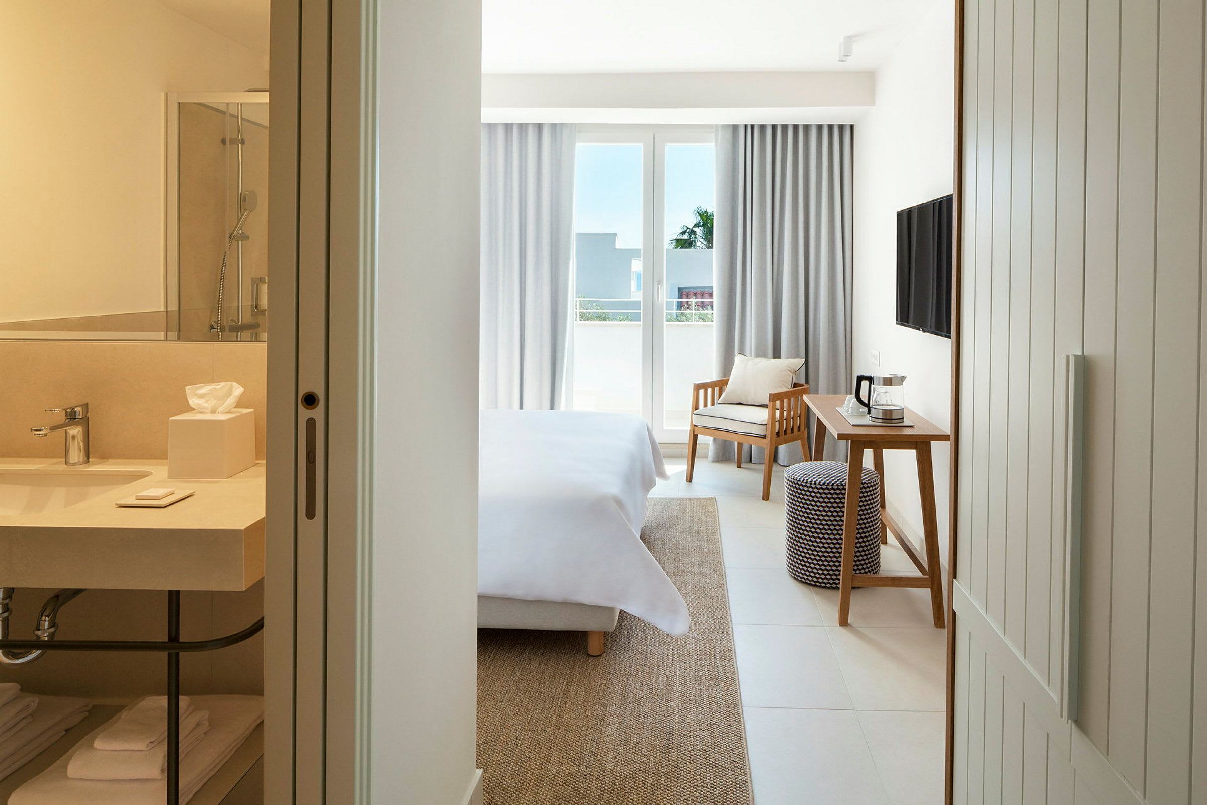 Et hotelværelse med en seng, et skrivebord, en stol og en balkon. I forgrunden ses et badeværelse. (AI-genereret tekst)