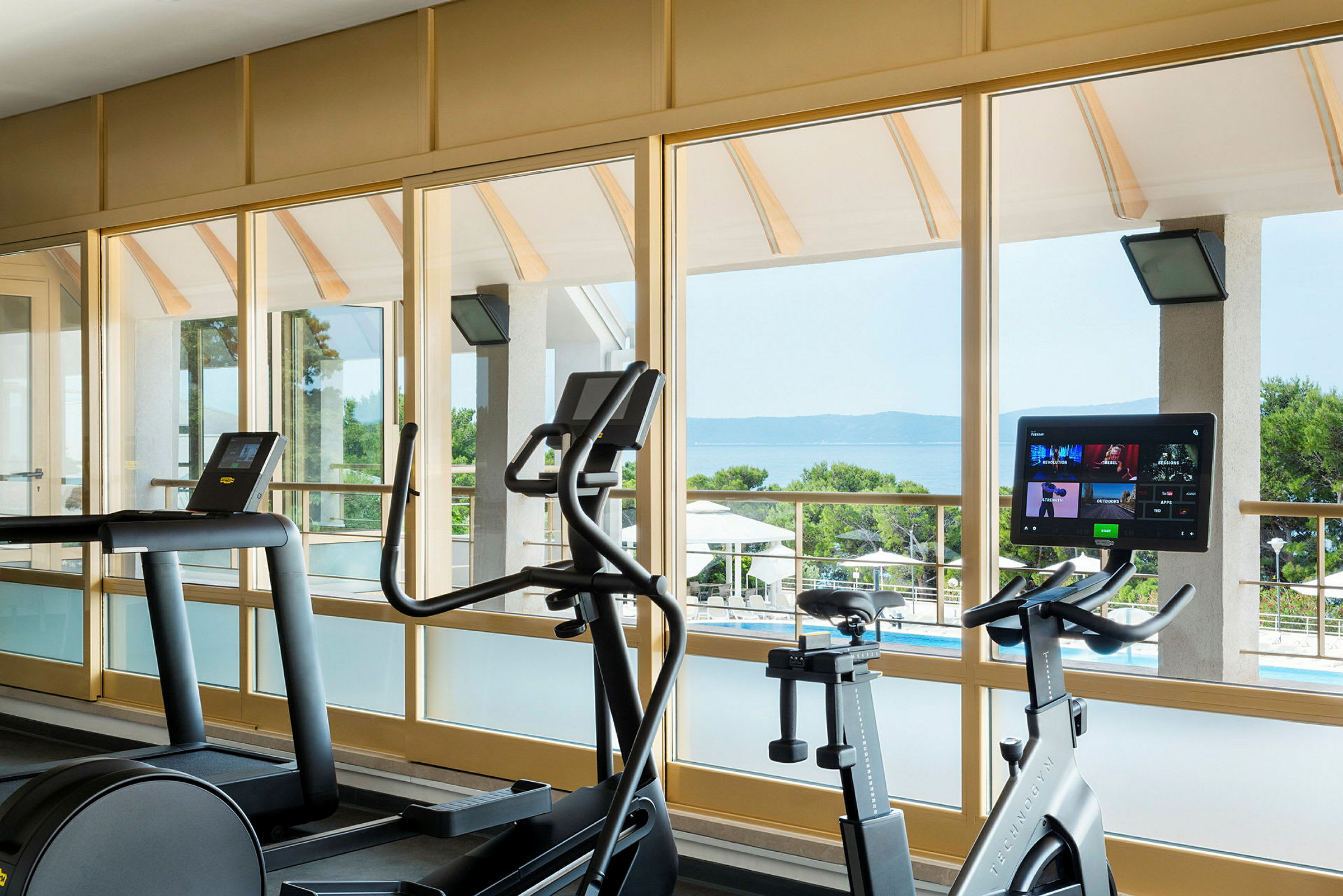 Fitnesscenter med træningsapparater foran store vinduer med udsigt over havet. (AI-genereret tekst)