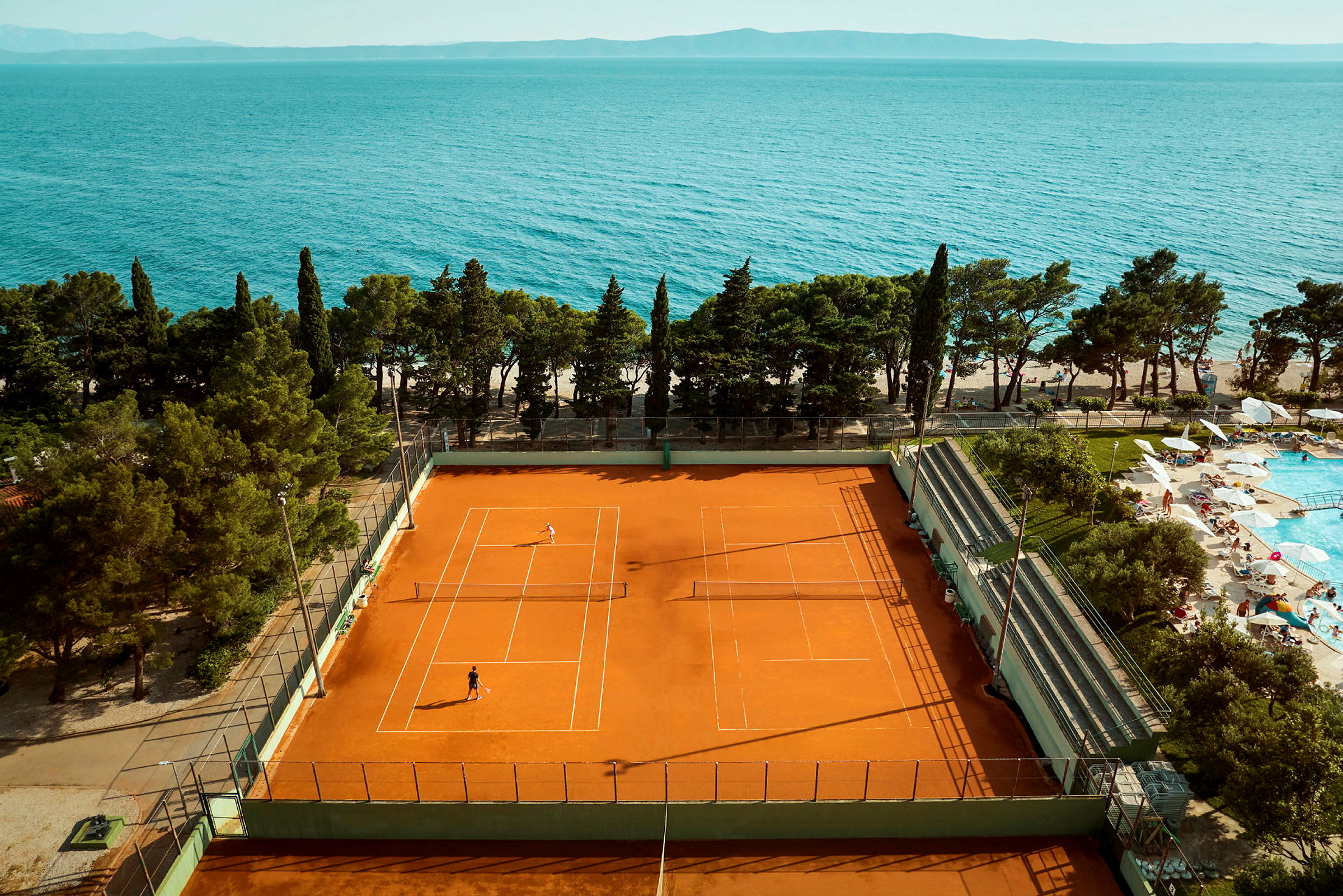 En tennisbane med spillere og et poolområde med liggestole ved siden af, samt hav i baggrunden. (AI-genereret tekst)