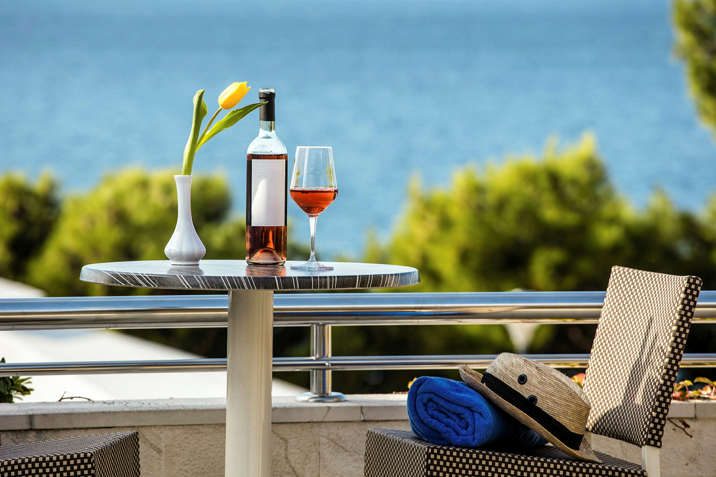 Et bord med en flaske og et glas rosévin samt en gul tulipan i vase, med udsigt over havet i baggrunden. (AI-genereret tekst)