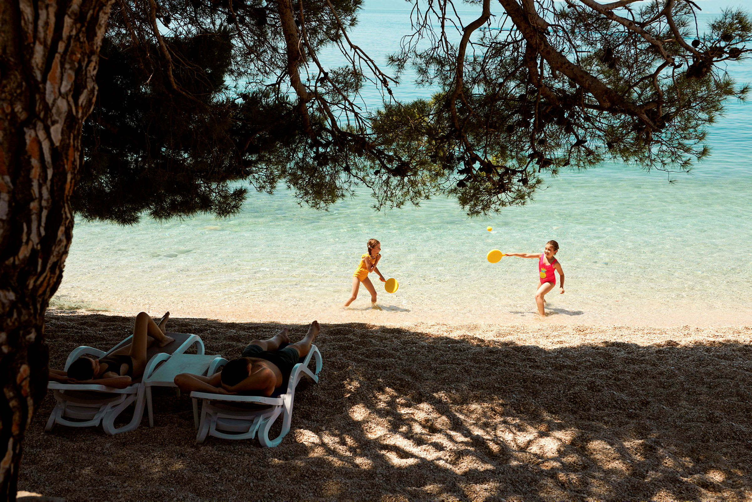 To børn leger med ketsjere i vandkanten på en strand. Flere personer ligger på solsenge under skyggefulde træer. (AI-genereret tekst)