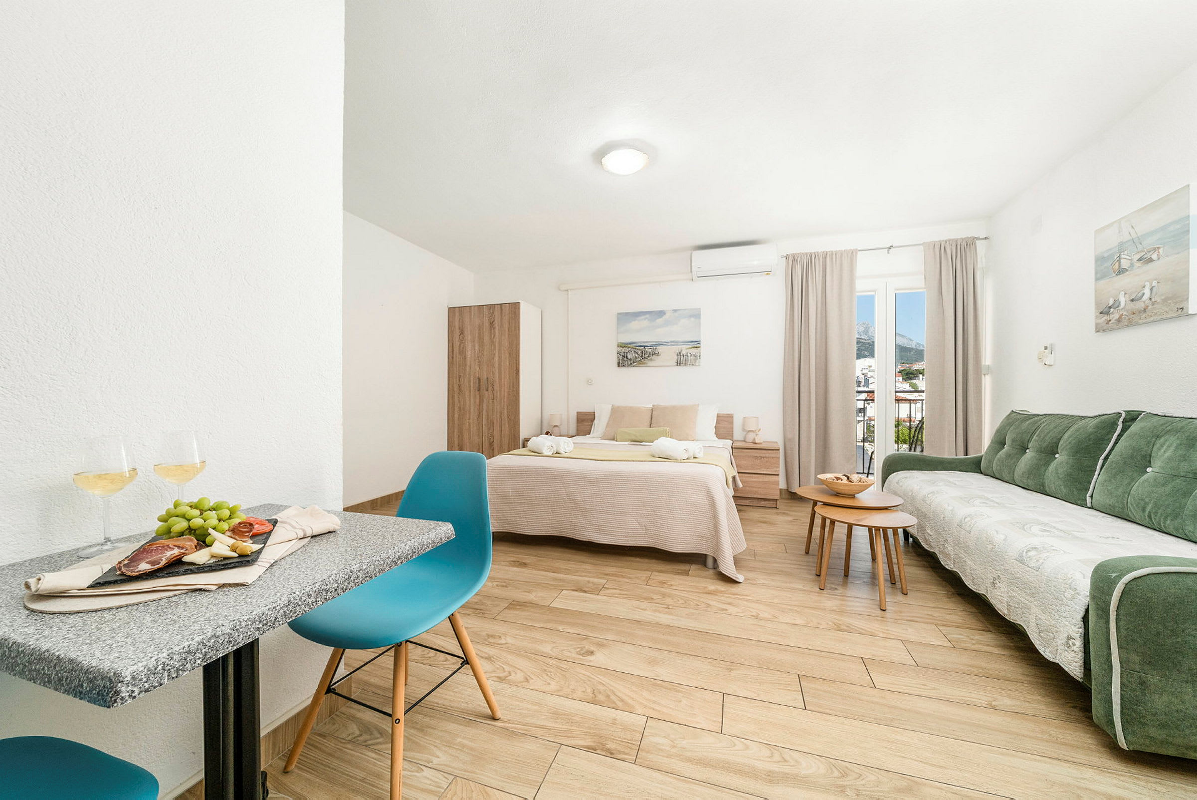 Et lyst og rummeligt værelse med en dobbeltseng, grøn sofa, bord og stole. Et sengebord står ved siden af sengen. Der er et vindue med gardiner. (AI-genereret tekst)