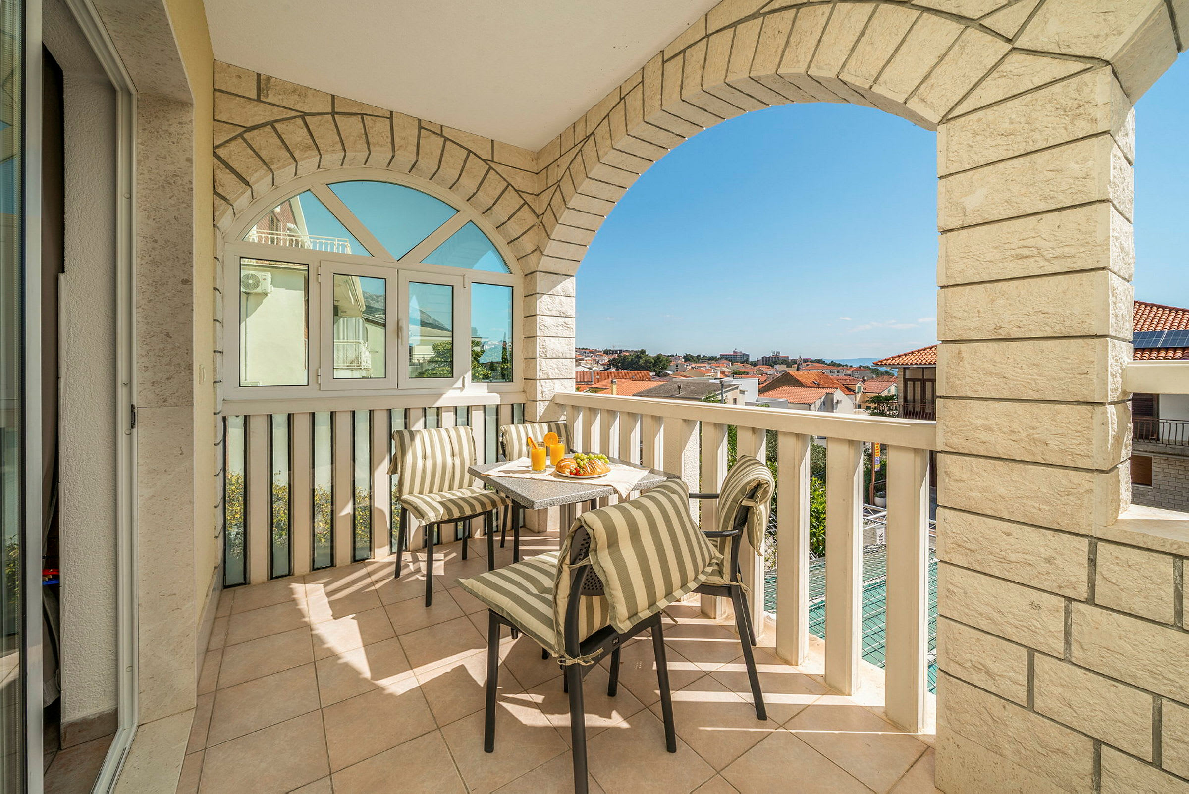 En balkon med bord og stole samt udsigt over omkringliggende hustage og en blå himmel. (AI-genereret tekst)