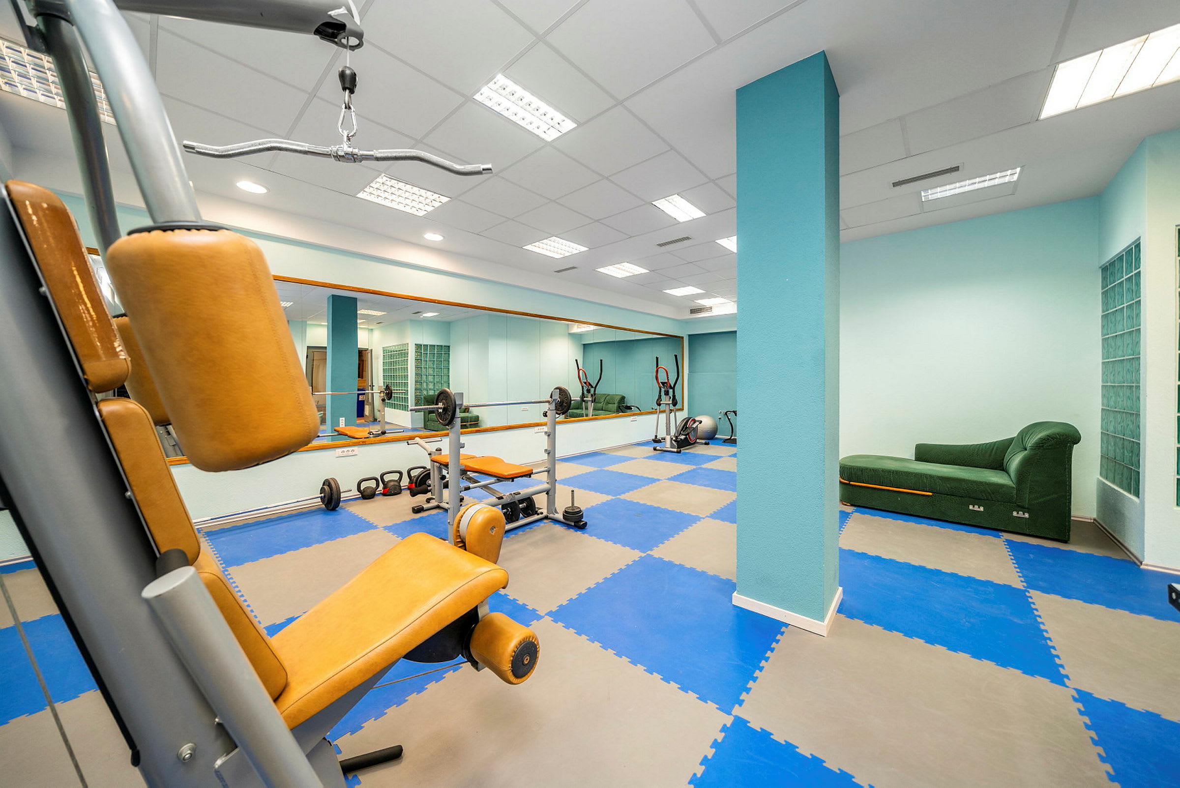 Et fitnessrum med træningsmaskiner, håndvægte og en grøn sofa. (AI-genereret tekst)