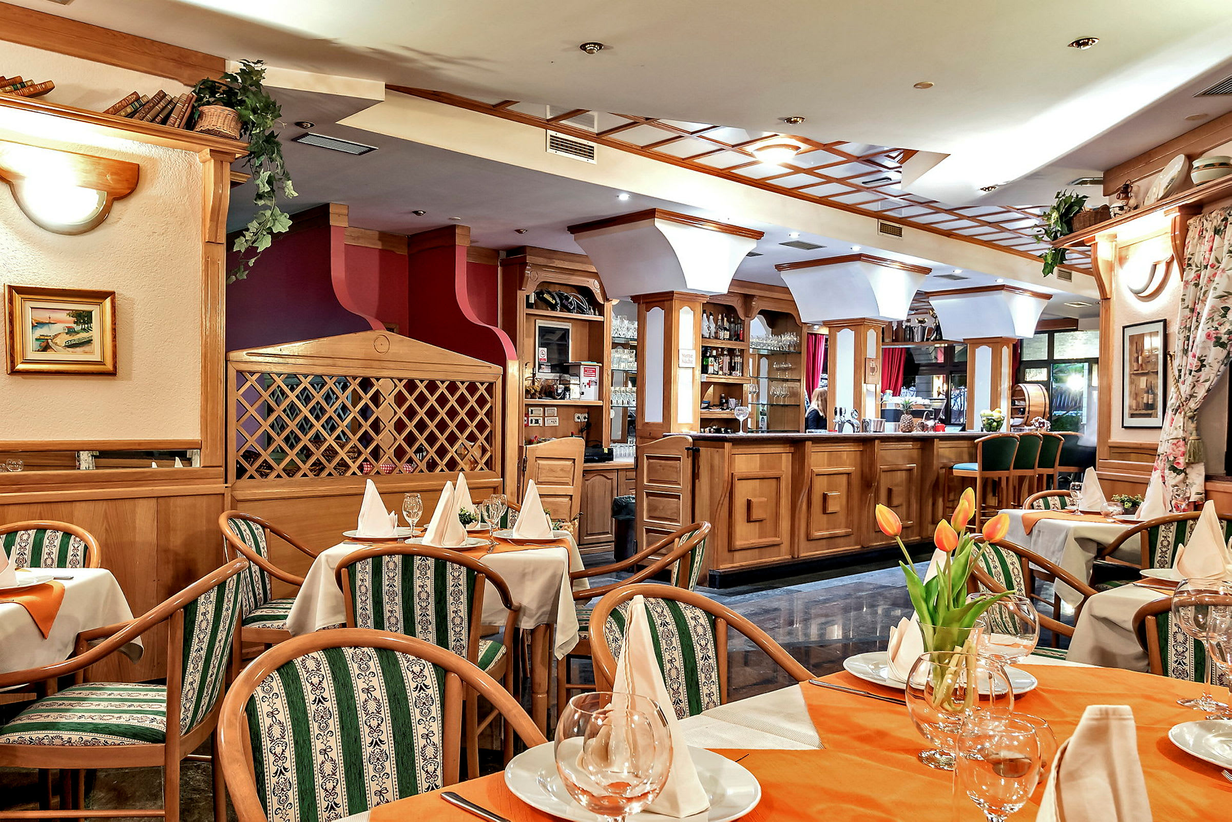 En indrettet restaurant med borde dækket med hvide servietter og glas. Stole med grønne mønstrede puder omgiver bordene, og en bardisk i træ er synlig i baggrunden. (AI-genereret tekst)