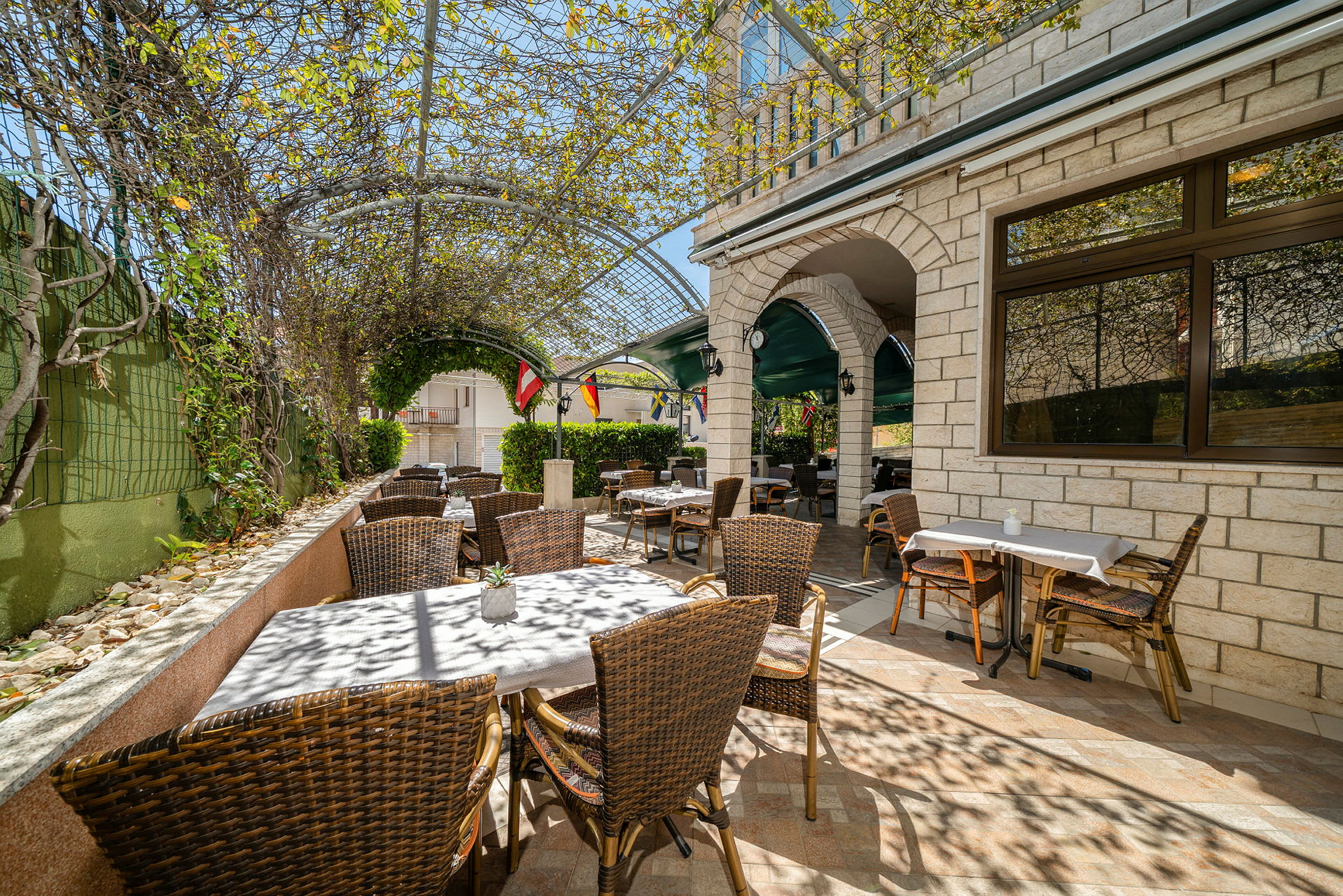 En udendørs café med kurvestole og borde under en pergola dækket af grønne planter. (AI-genereret tekst)