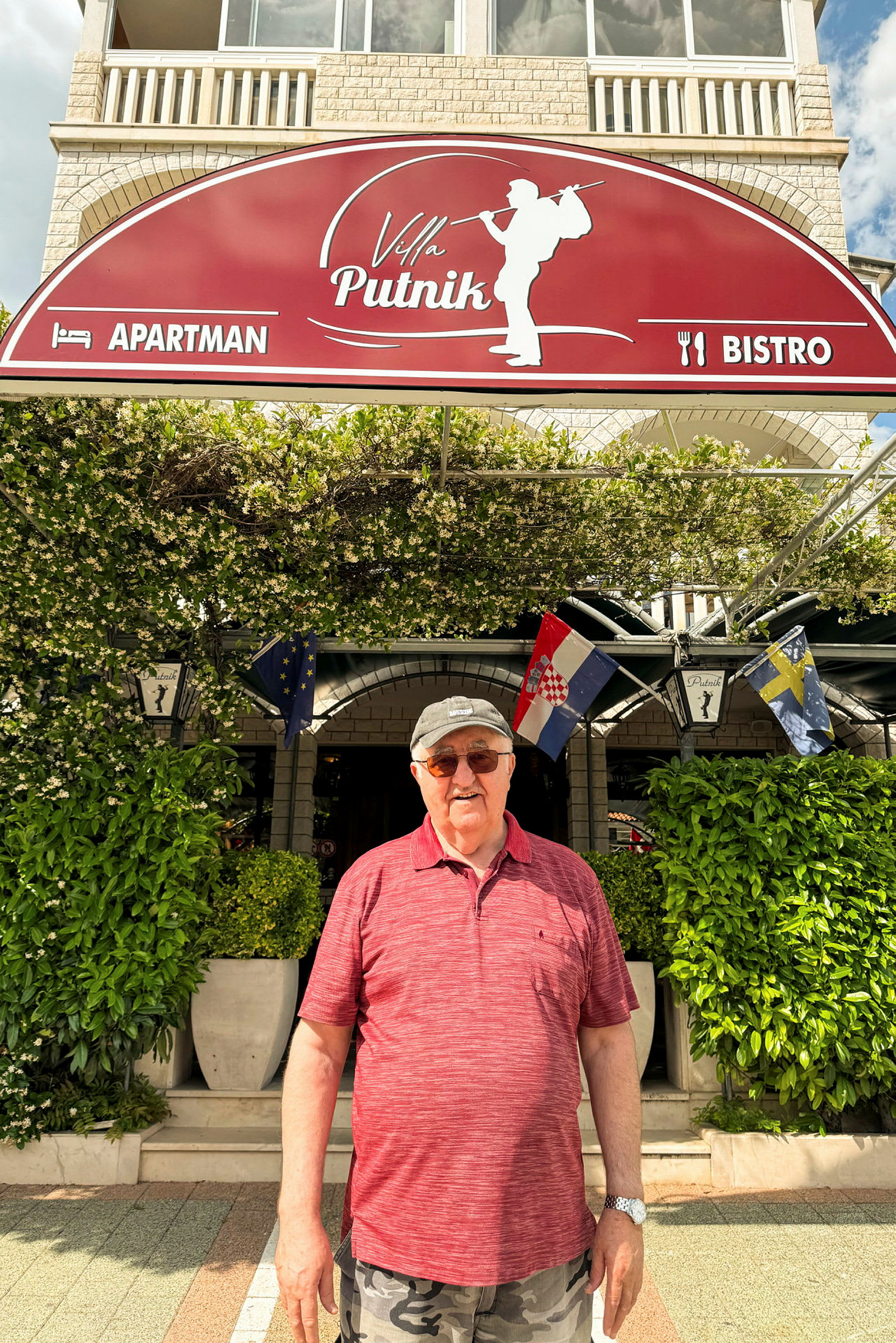 Et billede af en person, der står foran en bygning med et skilt med teksten "Villa Putnik" omgivet af grønne planter. (AI-genereret tekst)