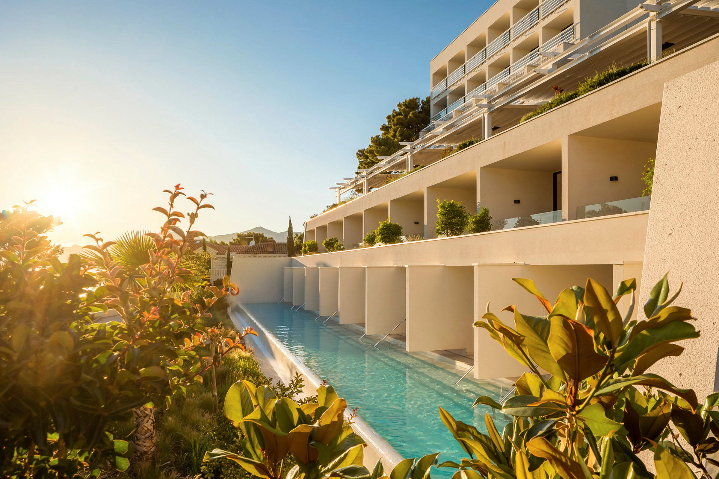 En hotelbygning med en langsgående pool foran, omgivet af planter og solopgang. (AI-genereret tekst)