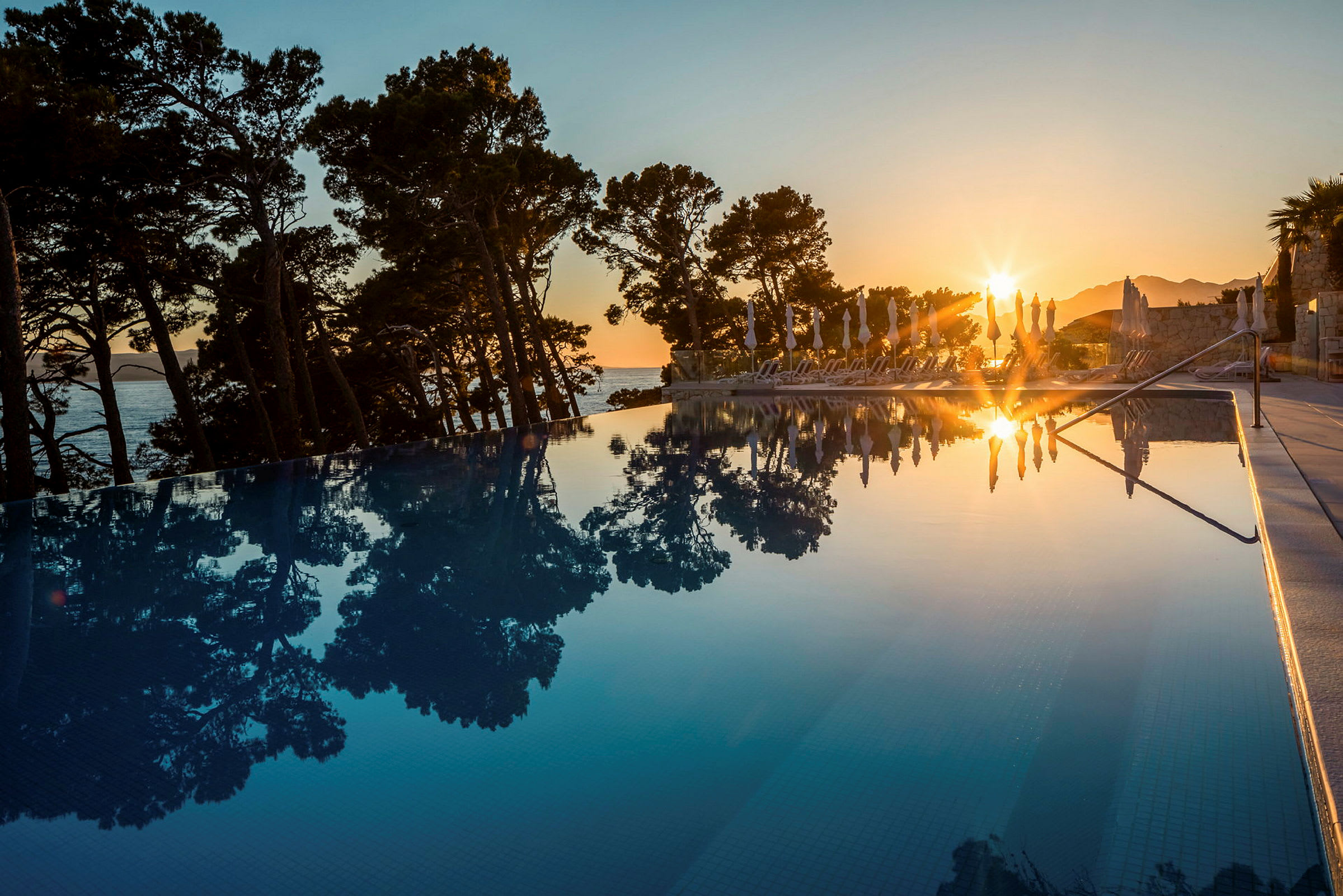 En solnedgang reflekteres i en pool, omgivet af fyrretræer og liggestole. (AI-genereret tekst)
