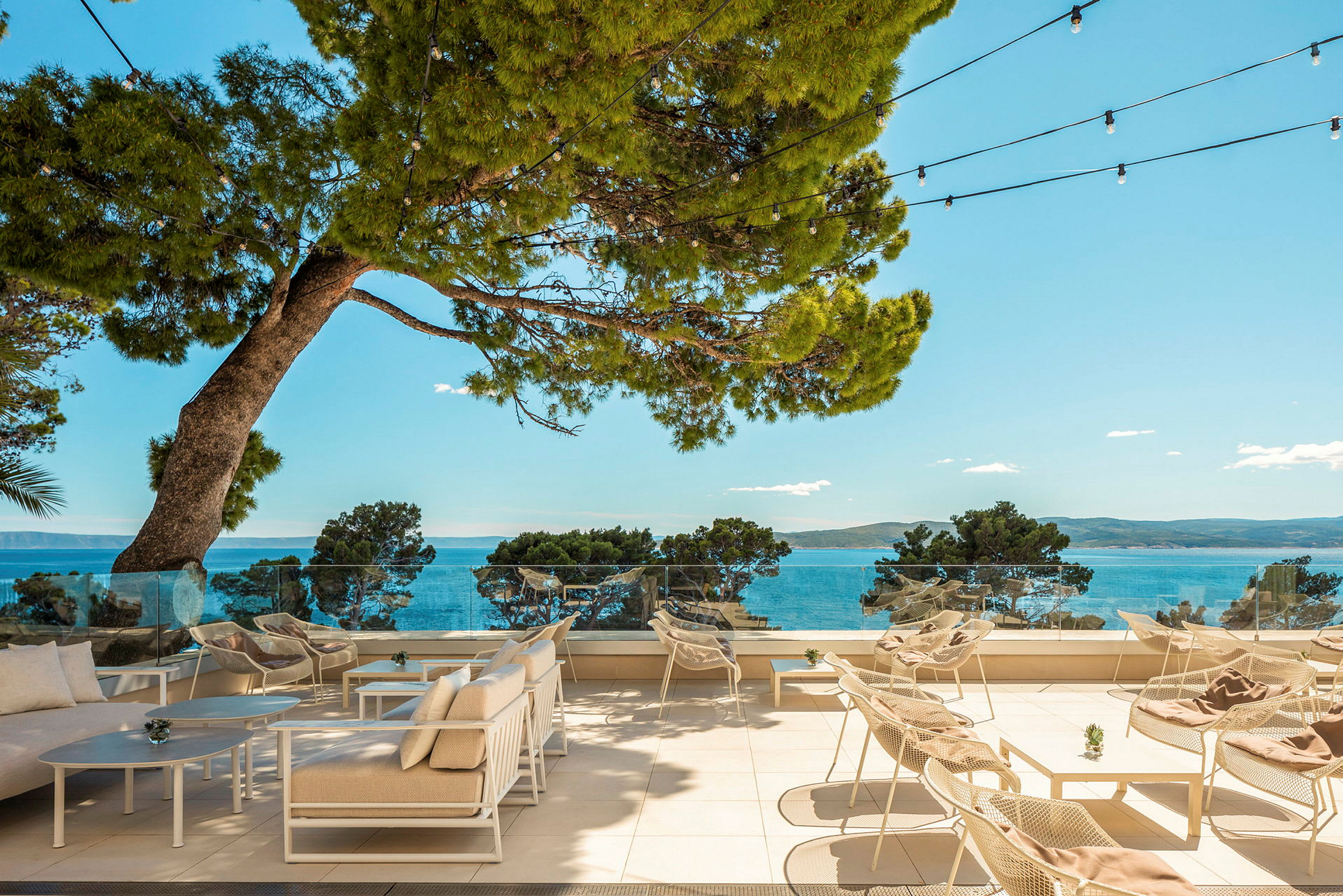 Udendørs terrasse med stole og borde, med udsigt til havet og de omkringliggende træer. (AI-genereret tekst)