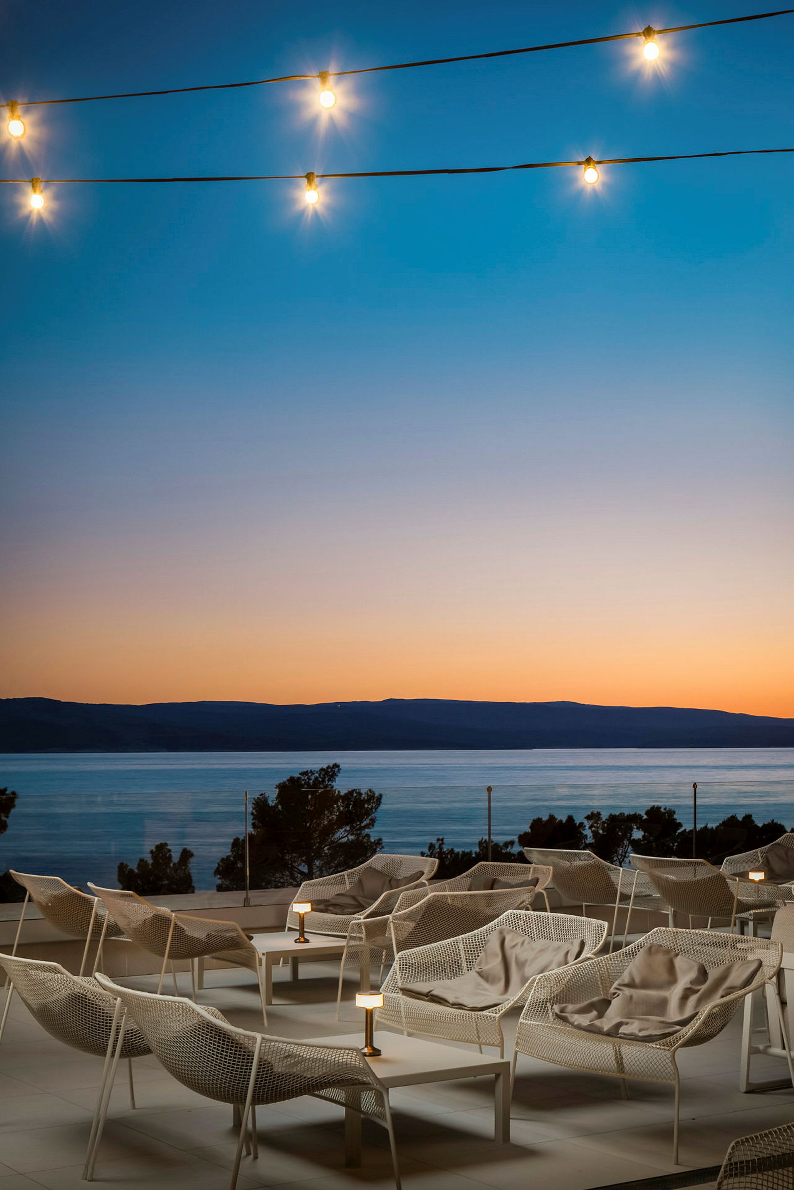 Hvide loungestole på en terrasse med udsigt over havet ved solnedgang, lys til dekoration. (AI-genereret tekst)