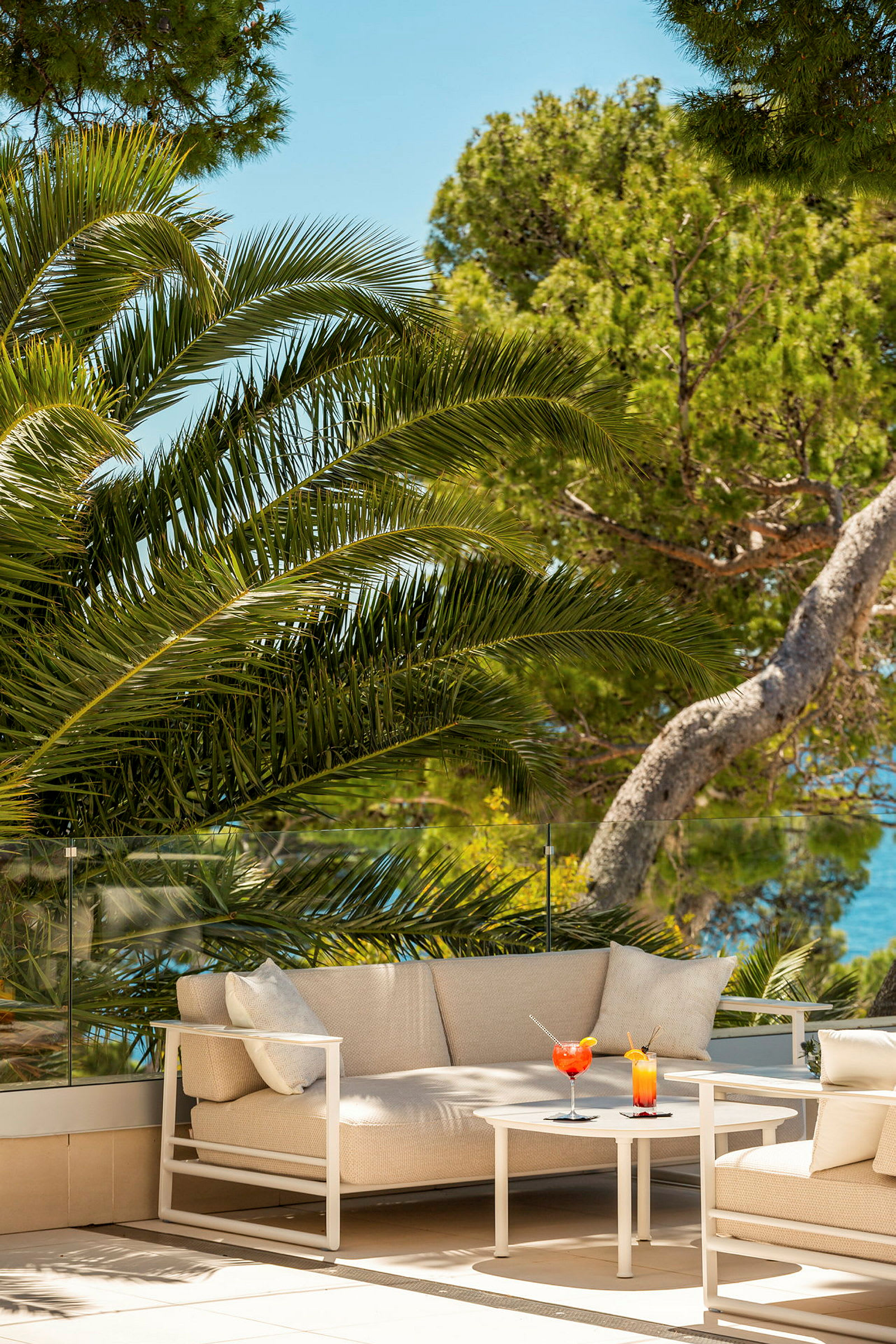 Lyse sofa og bord med to farverige drikkevarer udendørs omgivet af palmer og træer. (AI-genereret tekst)