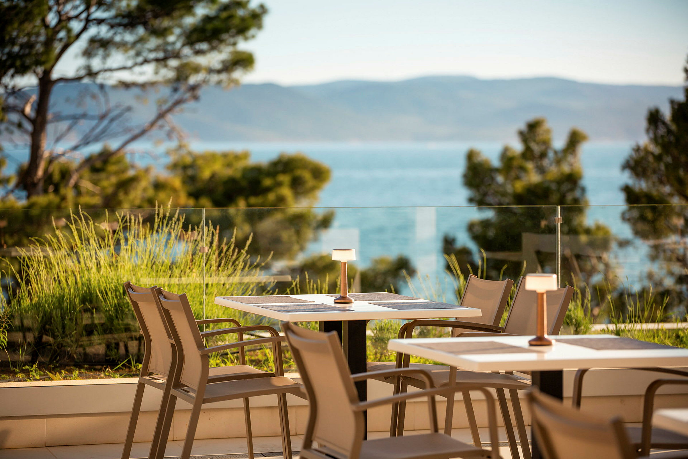 Udsigt over en terrasse med borde og stole, omgivet af grønt og med havet i baggrunden. (AI-genereret tekst)