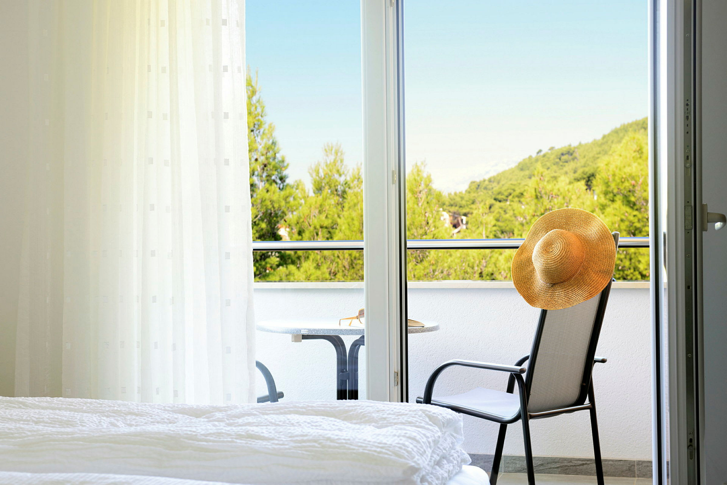 Et hotelværelse med udsigt til en balkon, hvor en stol med en hat og et bord står, samt frodige træer i baggrunden. (AI-genereret tekst)