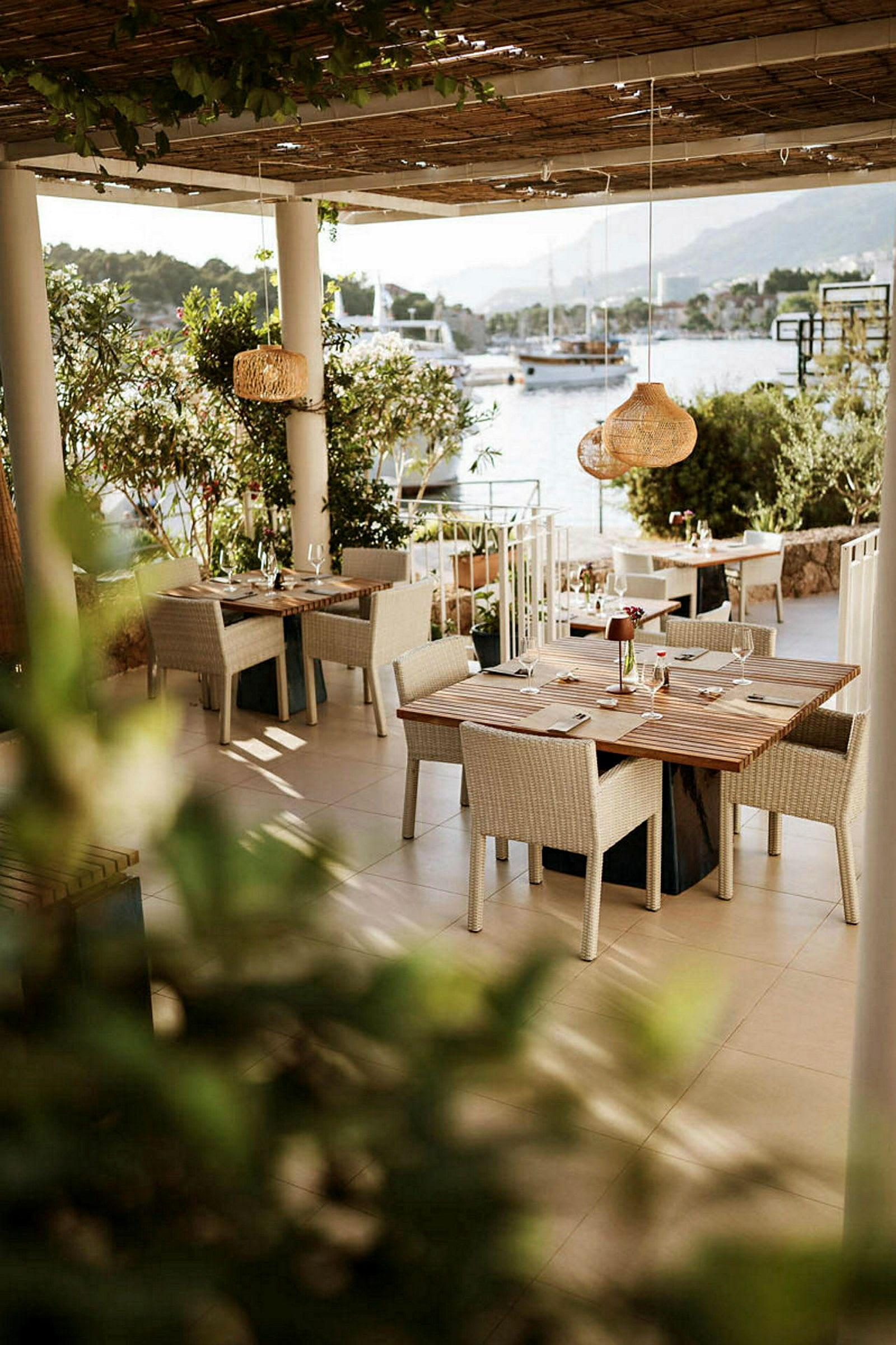 En udendørs restaurant med borde og stole under tag, nær vandet med både i baggrunden. (AI-genereret tekst)