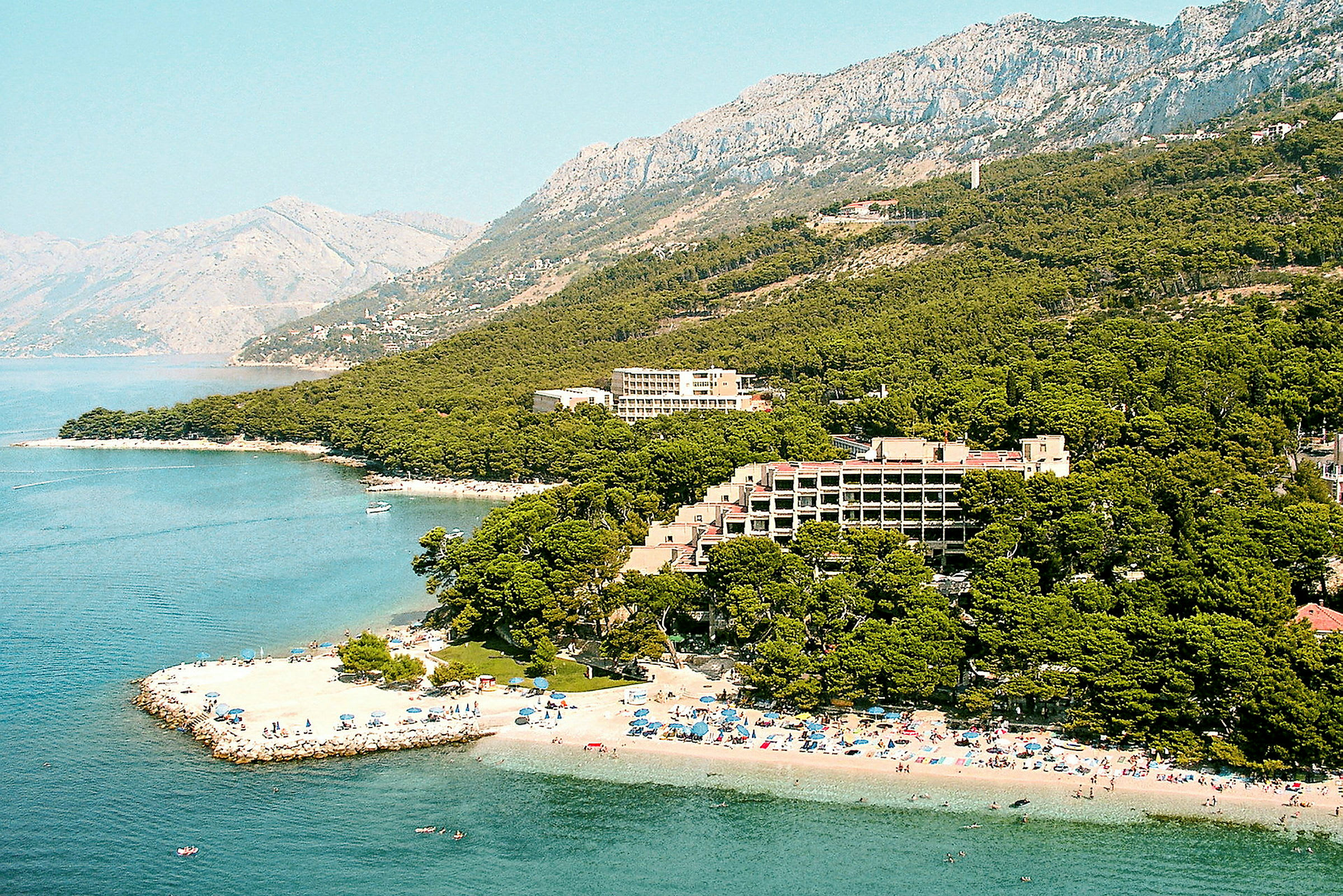 Strand, hotel og beplantning ved en kyst med bjerge i baggrunden. (AI-genereret tekst)