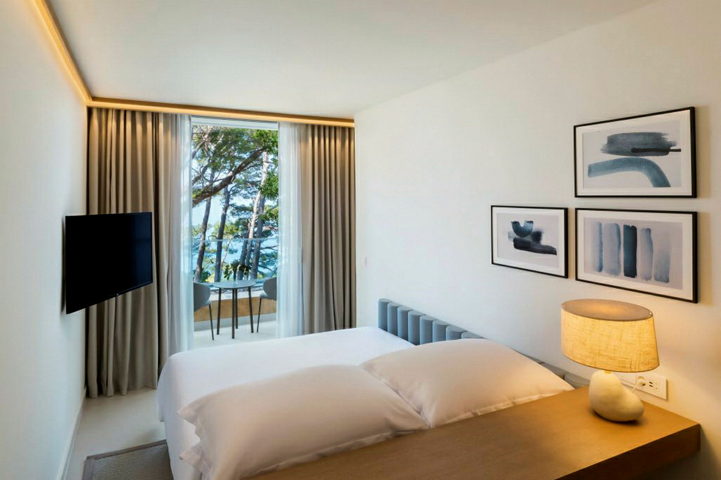 Et hotelværelse med en seng, en lampe på bordet, vægdekorationer og et vindue, der vender mod havet og træer. (AI-genereret tekst)