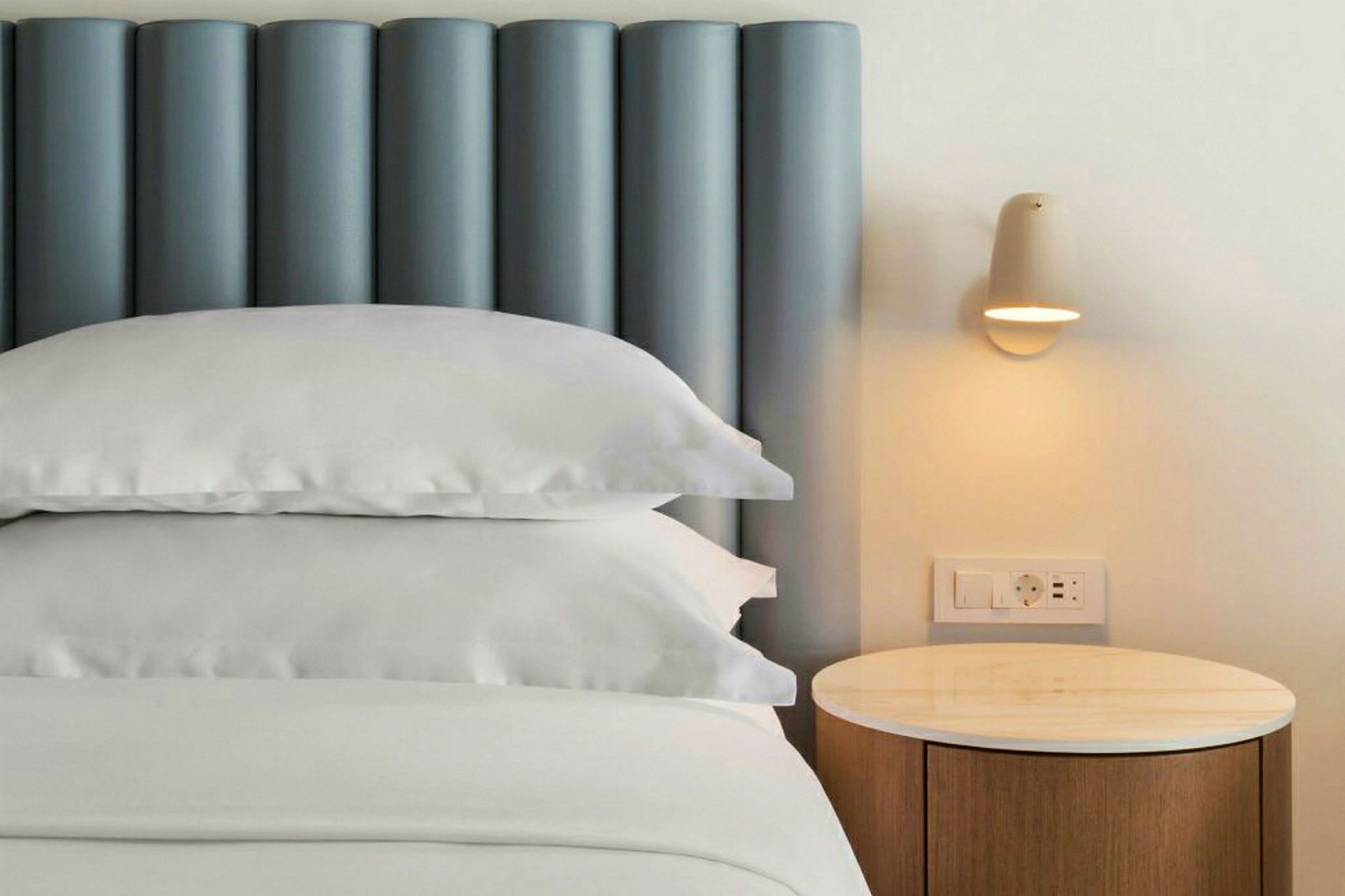 Nærbillede af en seng med en blå sengegavl, hvide puder og sengetøj, ved siden af et natbord med lampe. (AI-genereret tekst)