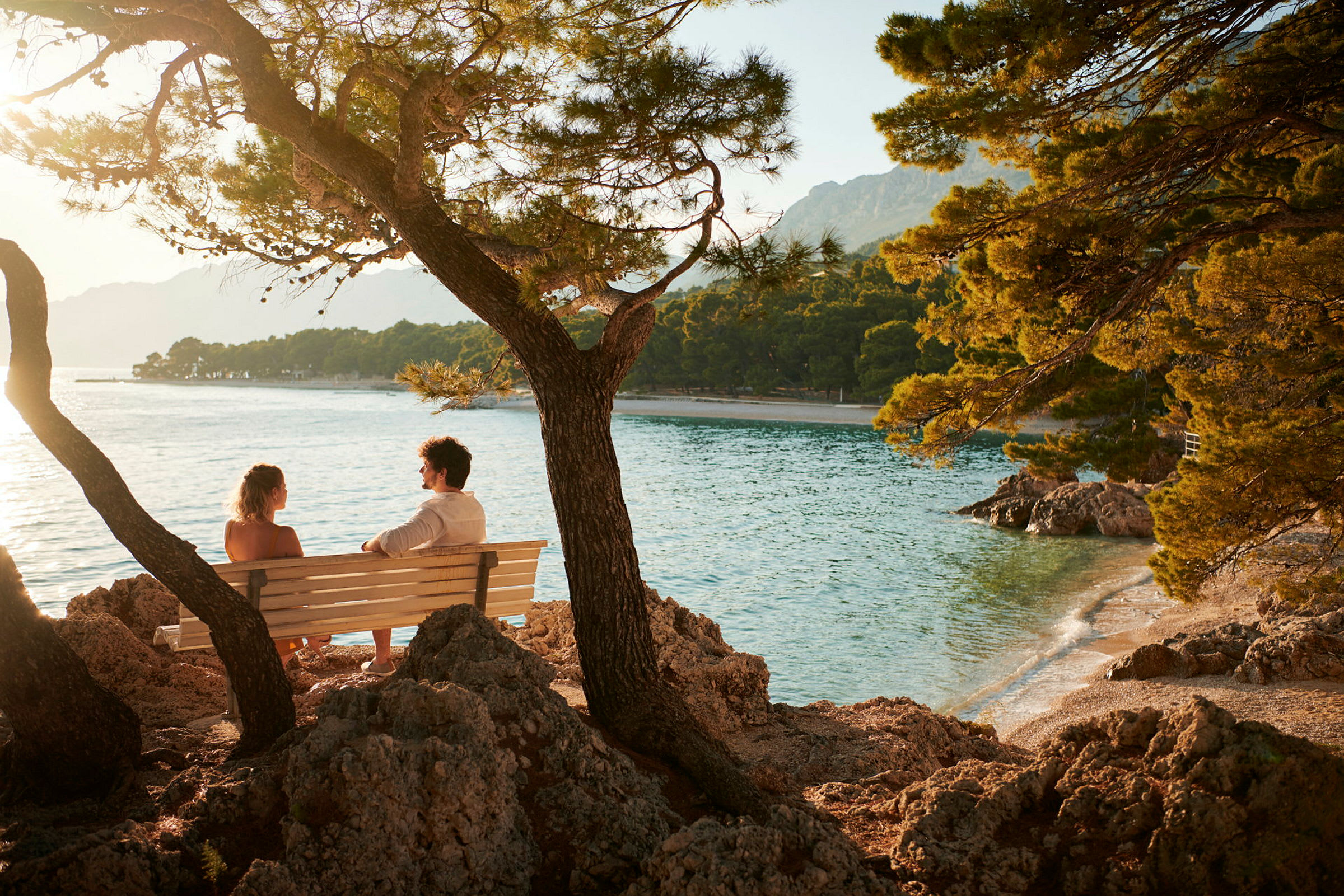 To personer sidder på en bænk ved kysten med udsigt over havet og træer. (AI-genereret tekst)