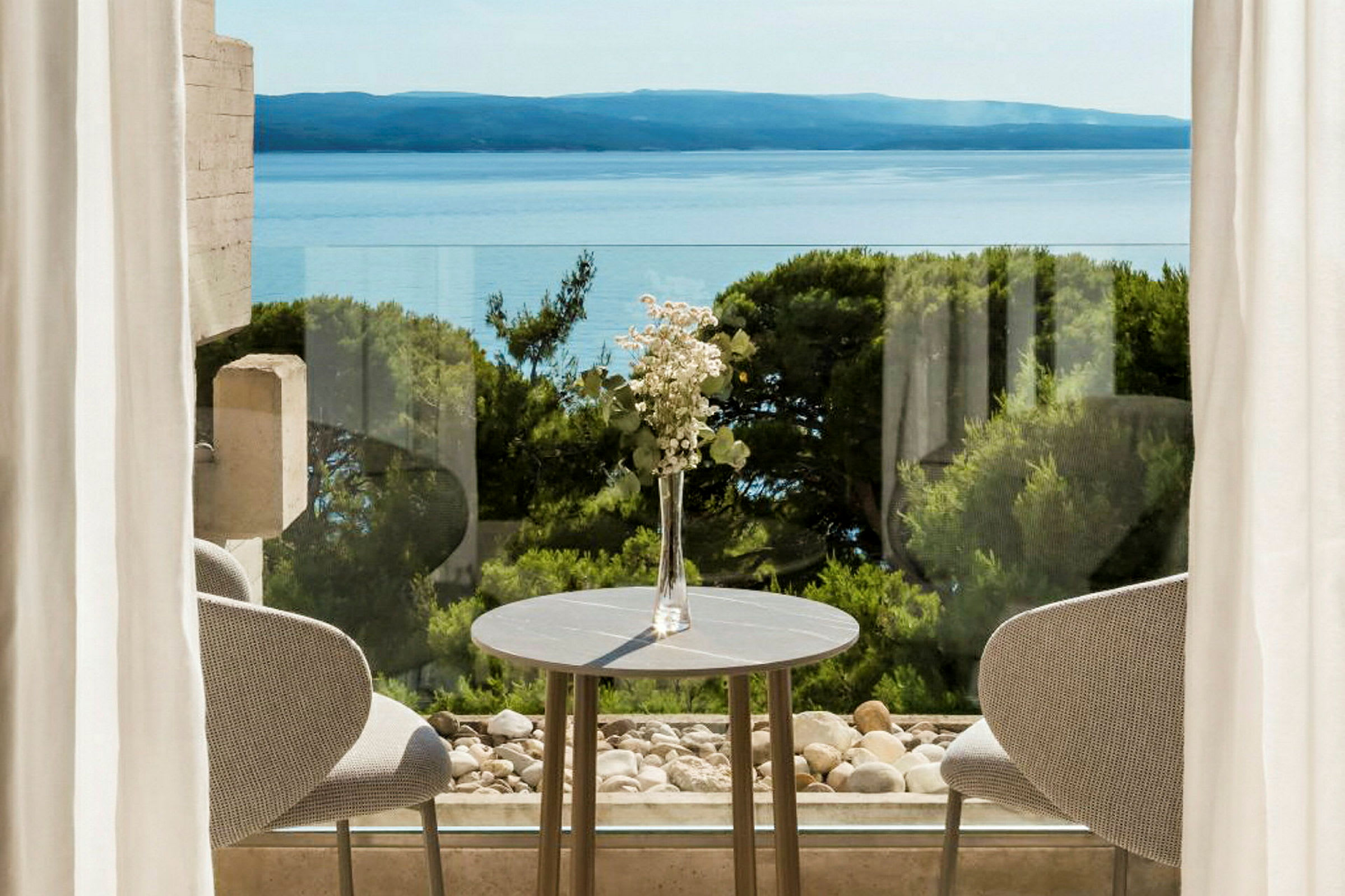 En borddekoration med blomster ved et bord foran vinduer med udsigt over havet og grønt. (AI-genereret tekst)