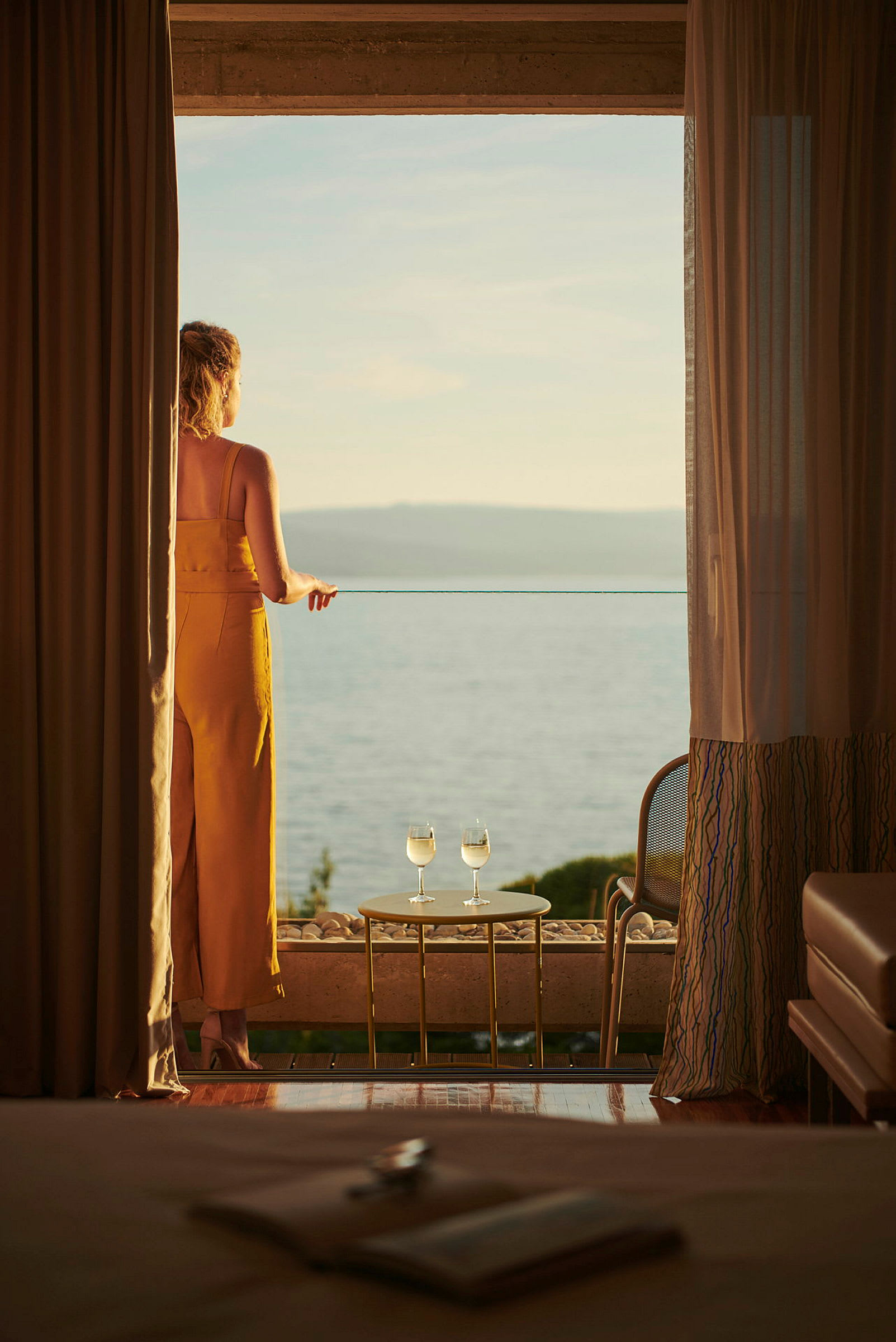 En kvinde står på en balkon og kigger ud over havet, med to glas hvidvin på et bord tæt ved hende. (AI-genereret tekst)